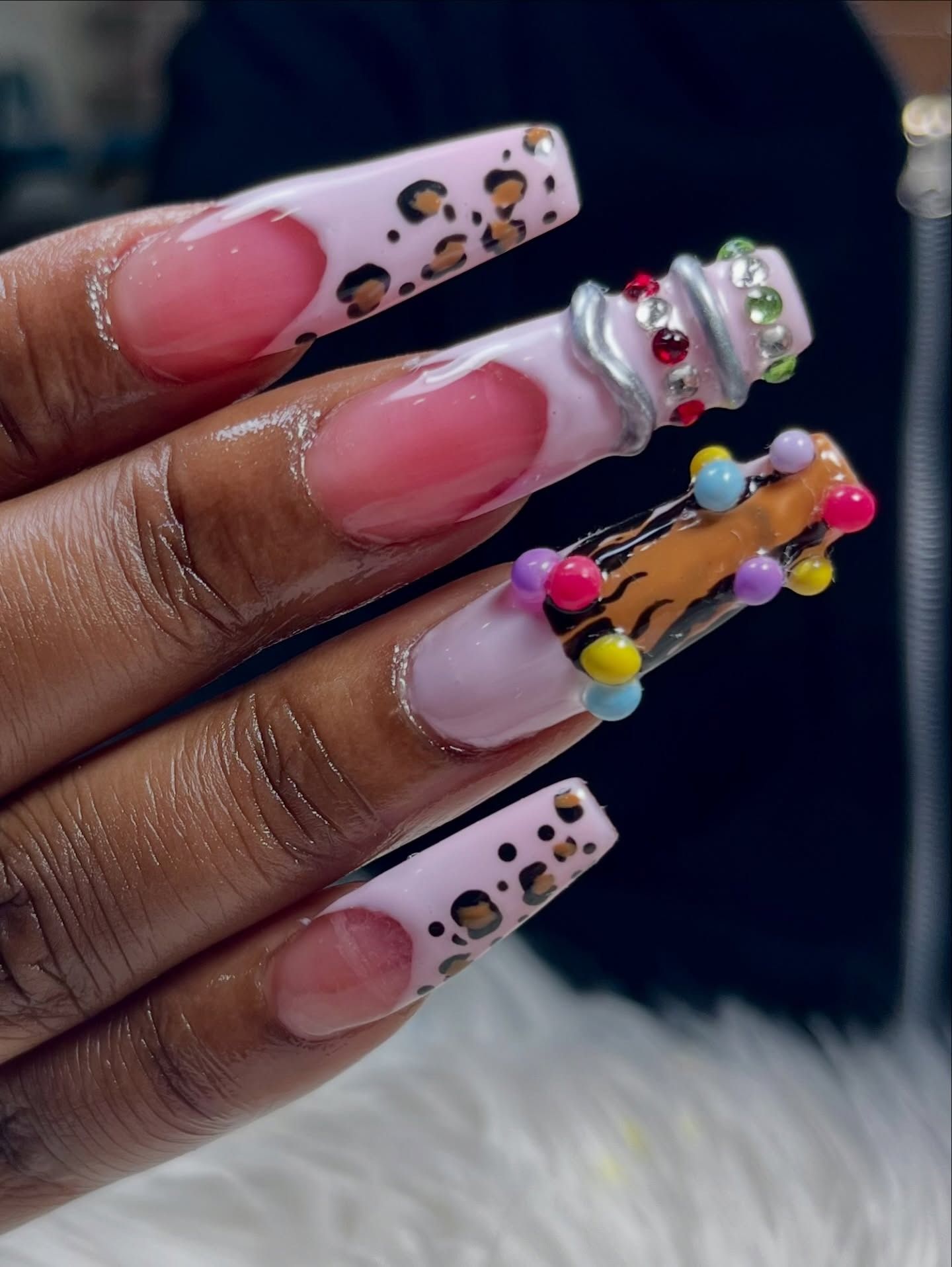 Hand with long pink and leopard-print acrylic nails, decorated with rhinestones and colorful charms