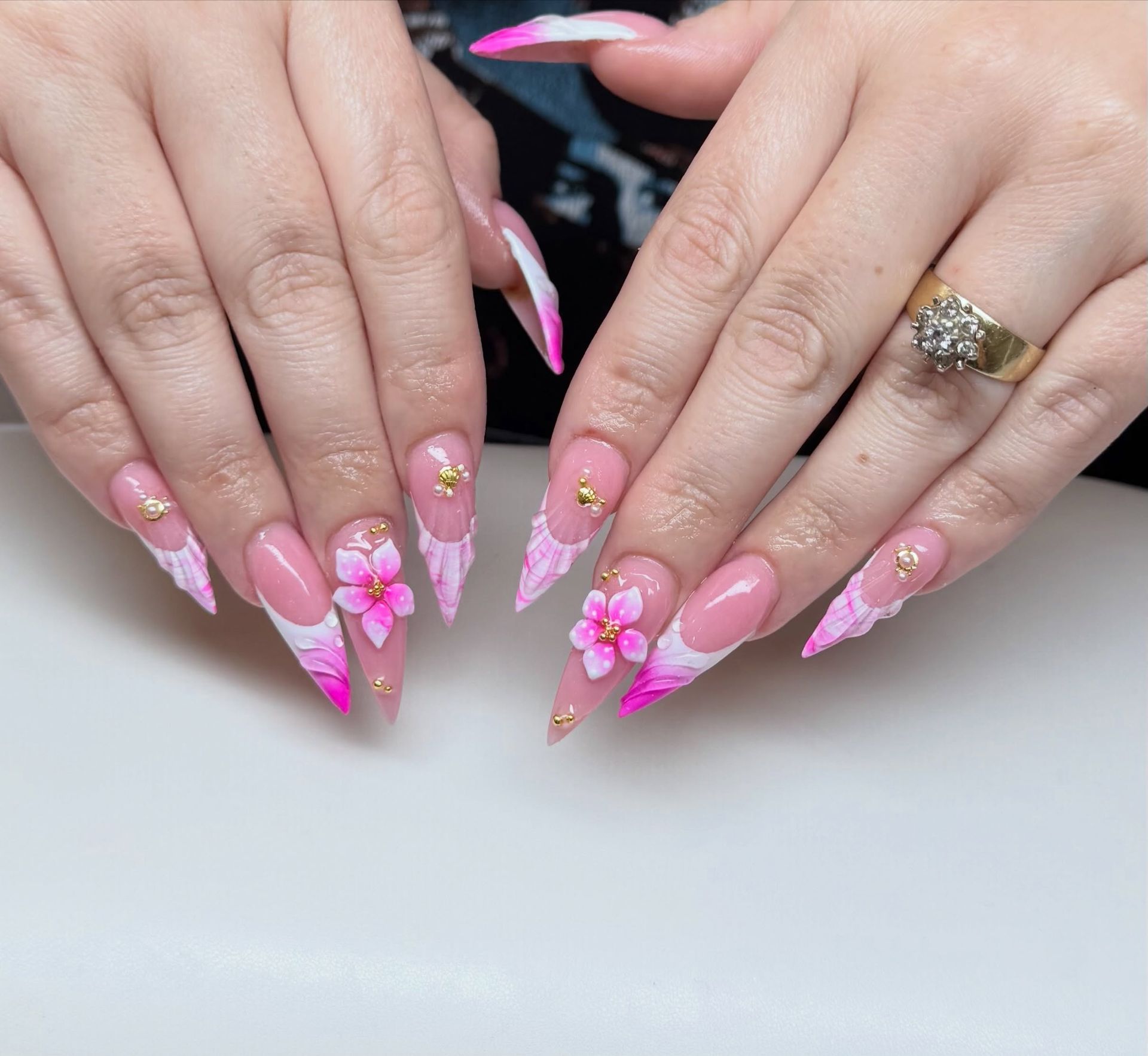 Hands with long pink ombré acrylic nails and 3D floral nail art, wearing a gold ring.