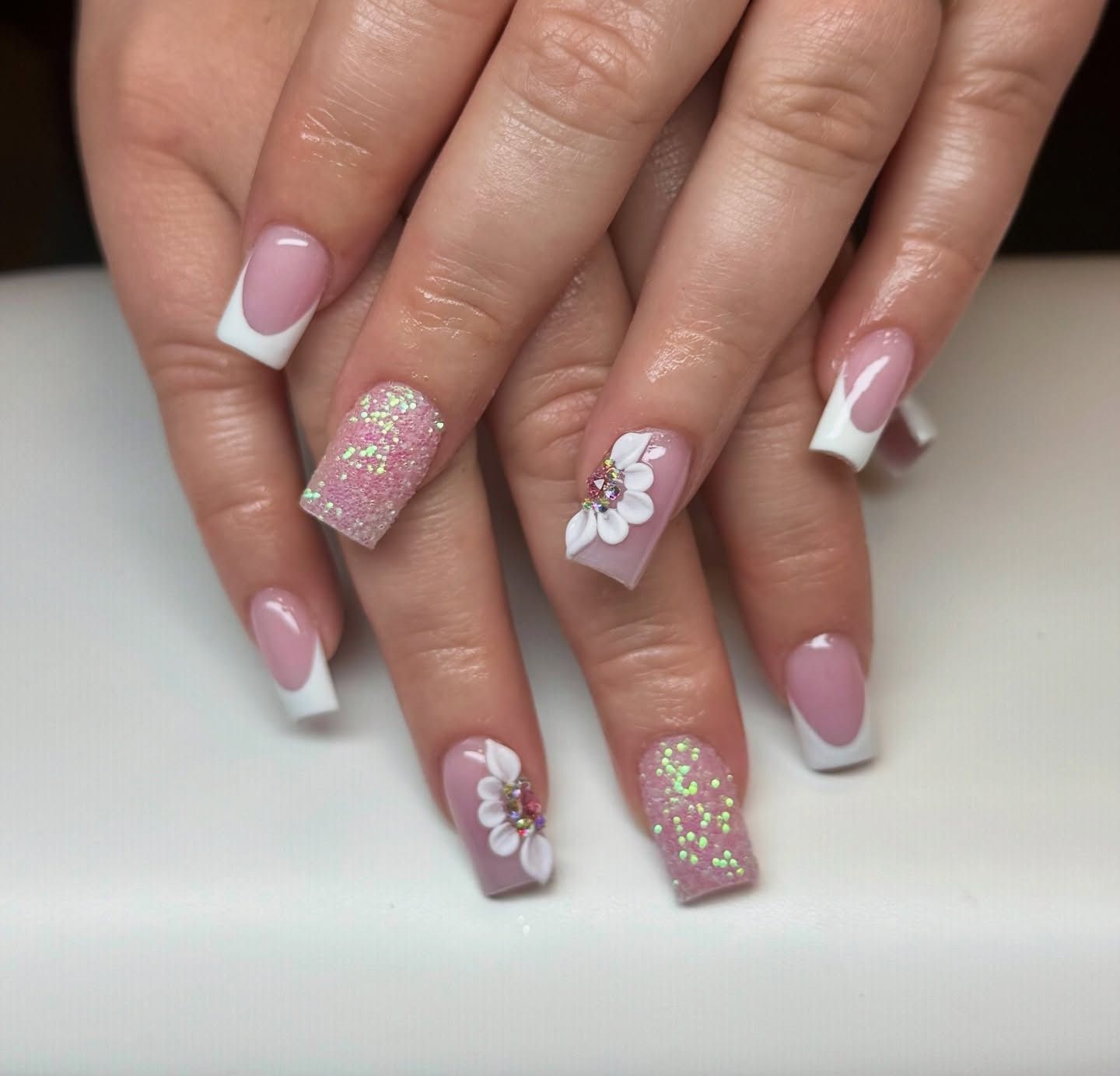 Hands with pink and white French manicure, glitter accents, and white flower nail art