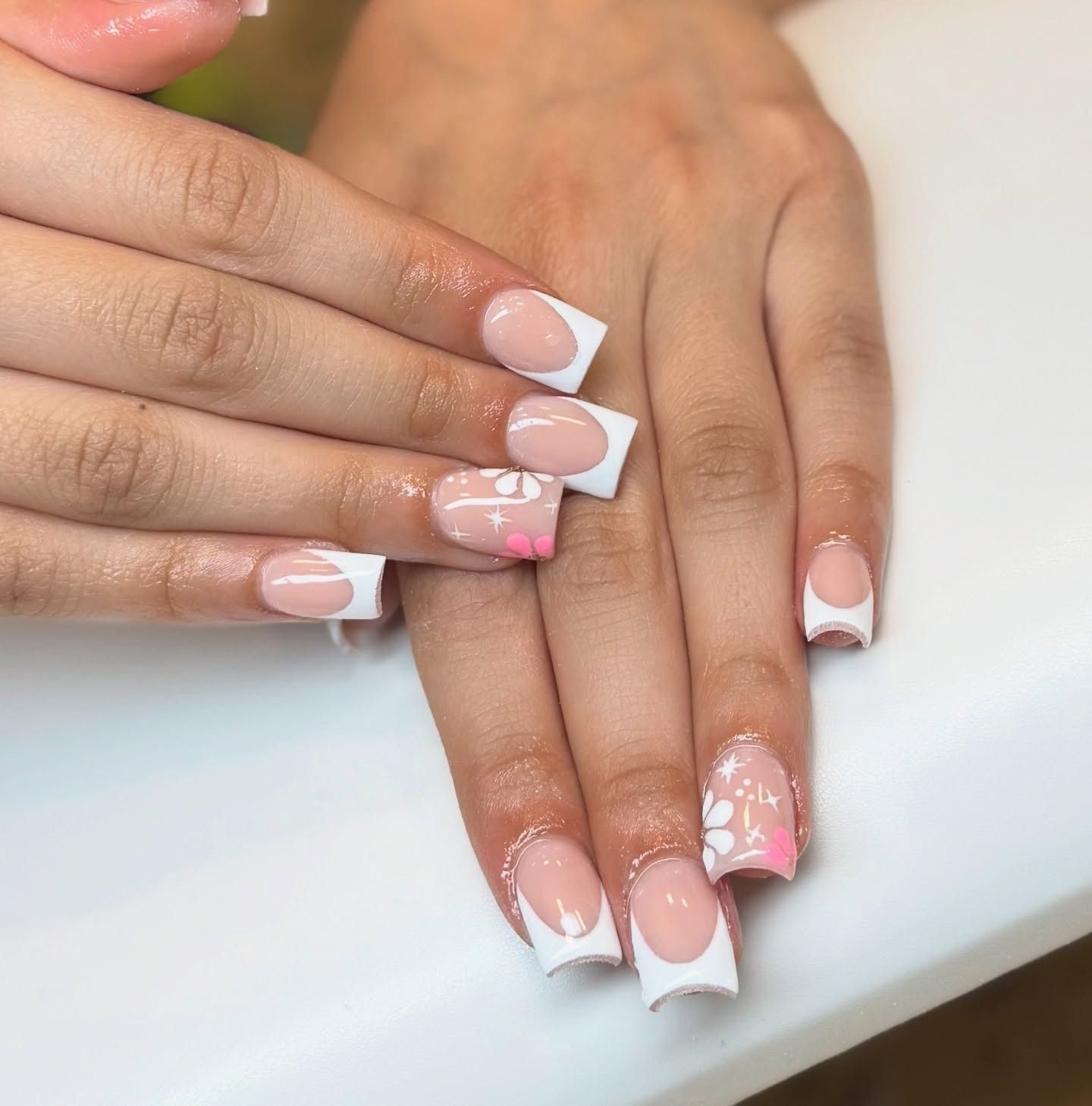 Hands with white French-tip nails and soft pink nail art resting together on a white surface.