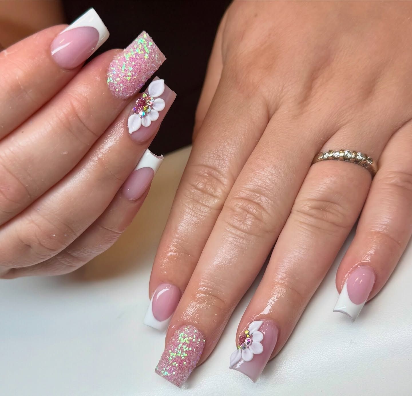 Pink glitter and white floral nail art on manicured hands with French tips and a ring