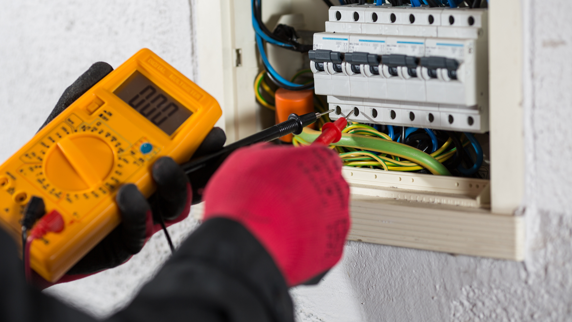 Electrician testing exposed wiring in a wall panel with a yellow multimeter and gloved hand.