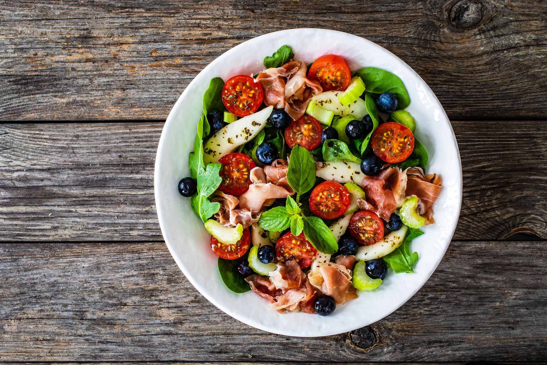 Colorful salad with cherry tomatoes, avocado, berries, and fresh basil in a white bowl on rustic wood table