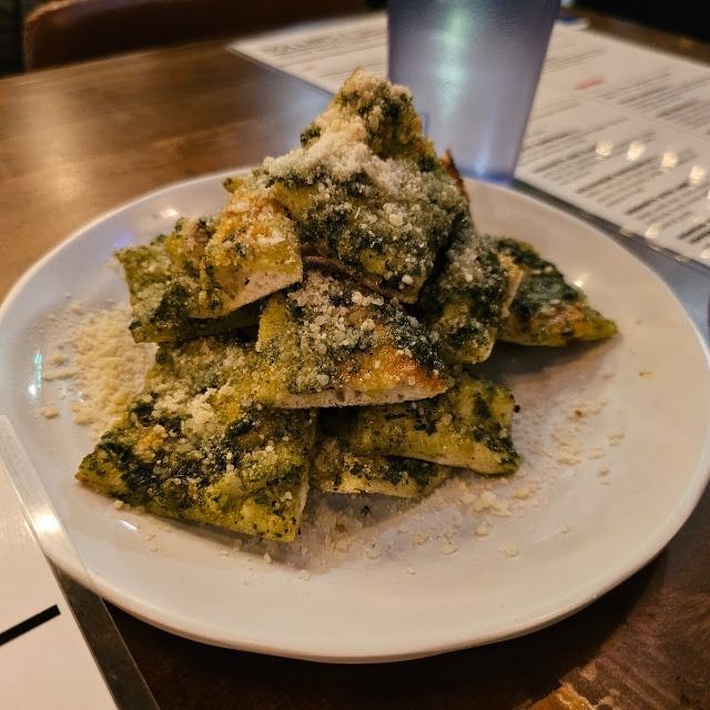 Plate of pasta covered in green sauce and grated cheese on a table with a blurred drink behind it