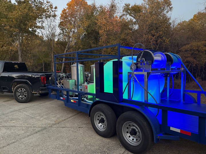 A black pickup truck tows a blue flatbed trailer featuring large water tanks and equipment, parked on a paved lot.