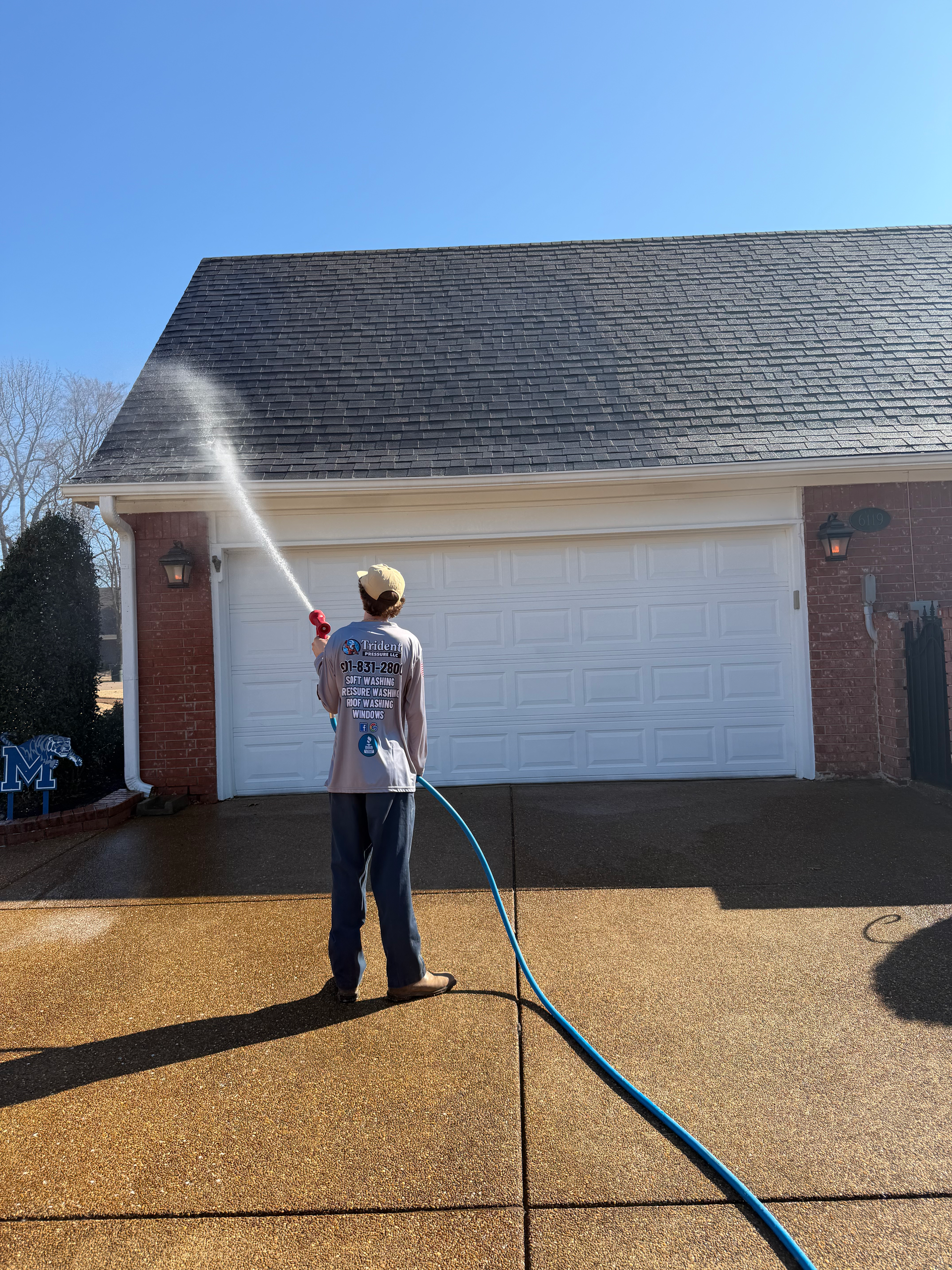 A person sprays water from a hose onto a shingled roof above a white garage door.