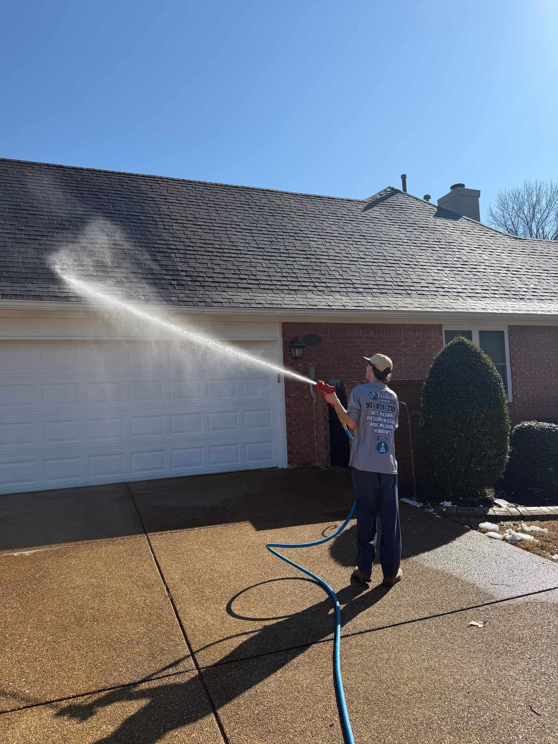 A person in a gray t-shirt uses a hose to spray water onto a dark-shingled roof above a white garage door.