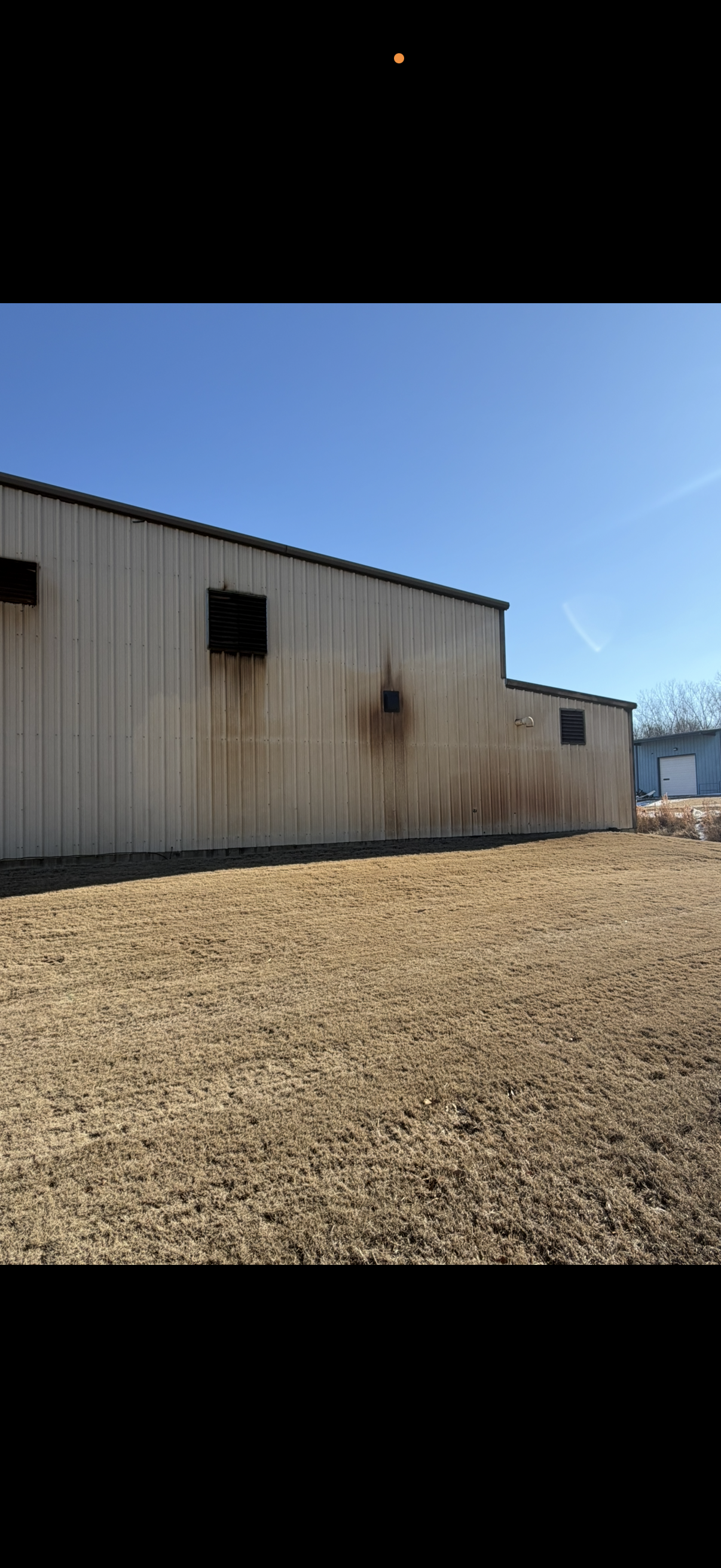 A tan metal building exterior with rust stains beneath several small windows against a clear blue sky.