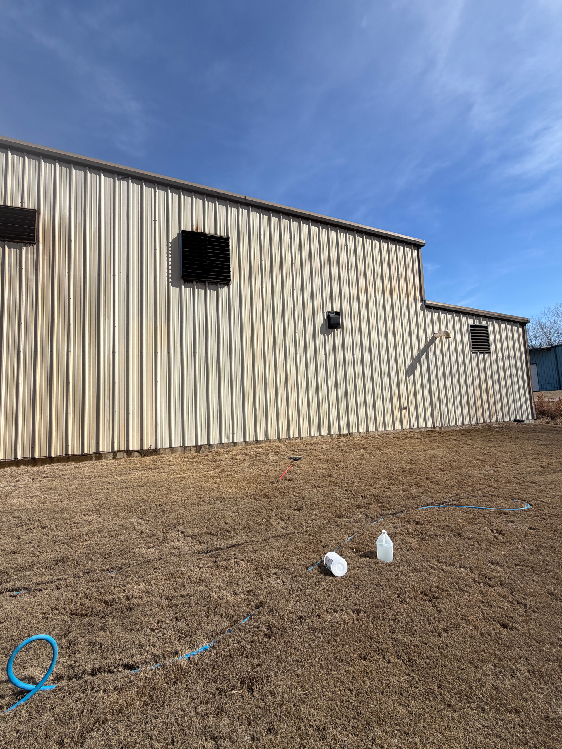 A side view of a beige metal-sided building under a clear blue sky, with two plastic bottles sitting on the dry grass.