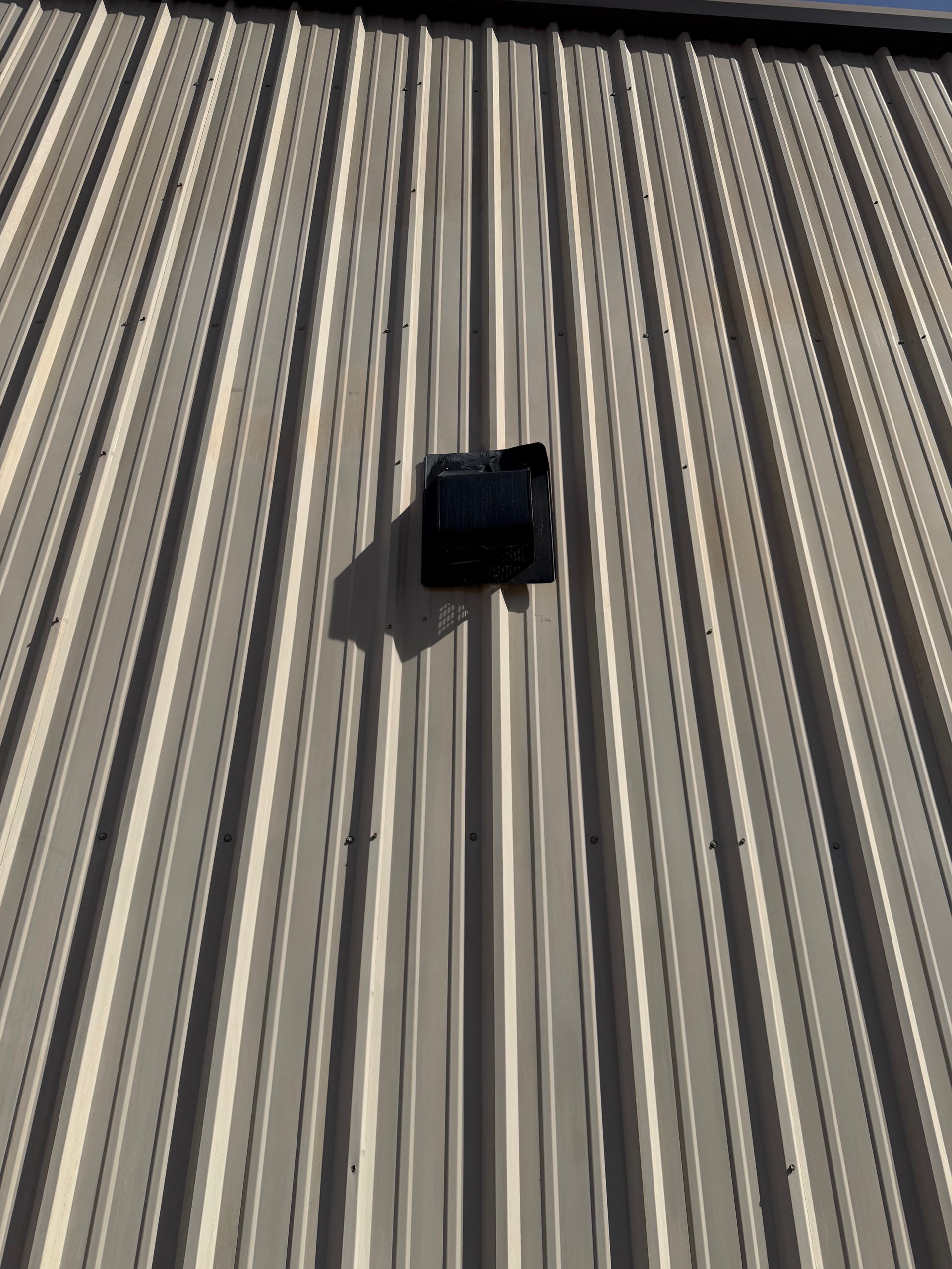 A black rectangular wall vent mounted on a light-colored, ribbed metal siding exterior wall.
