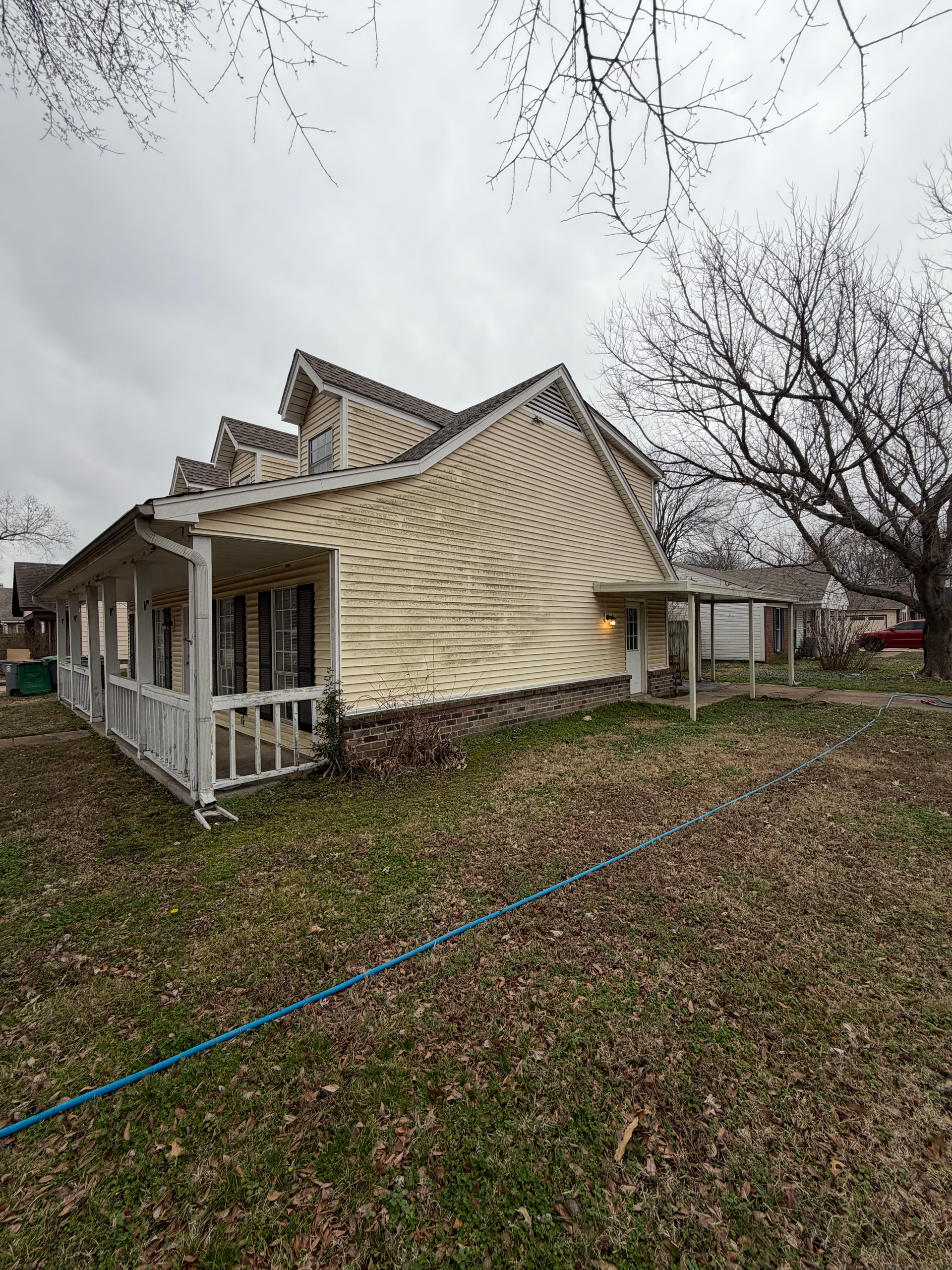 A side view of a house with pale yellow siding, a white porch, and a roof, surrounded by trees and a brown grassy yard.