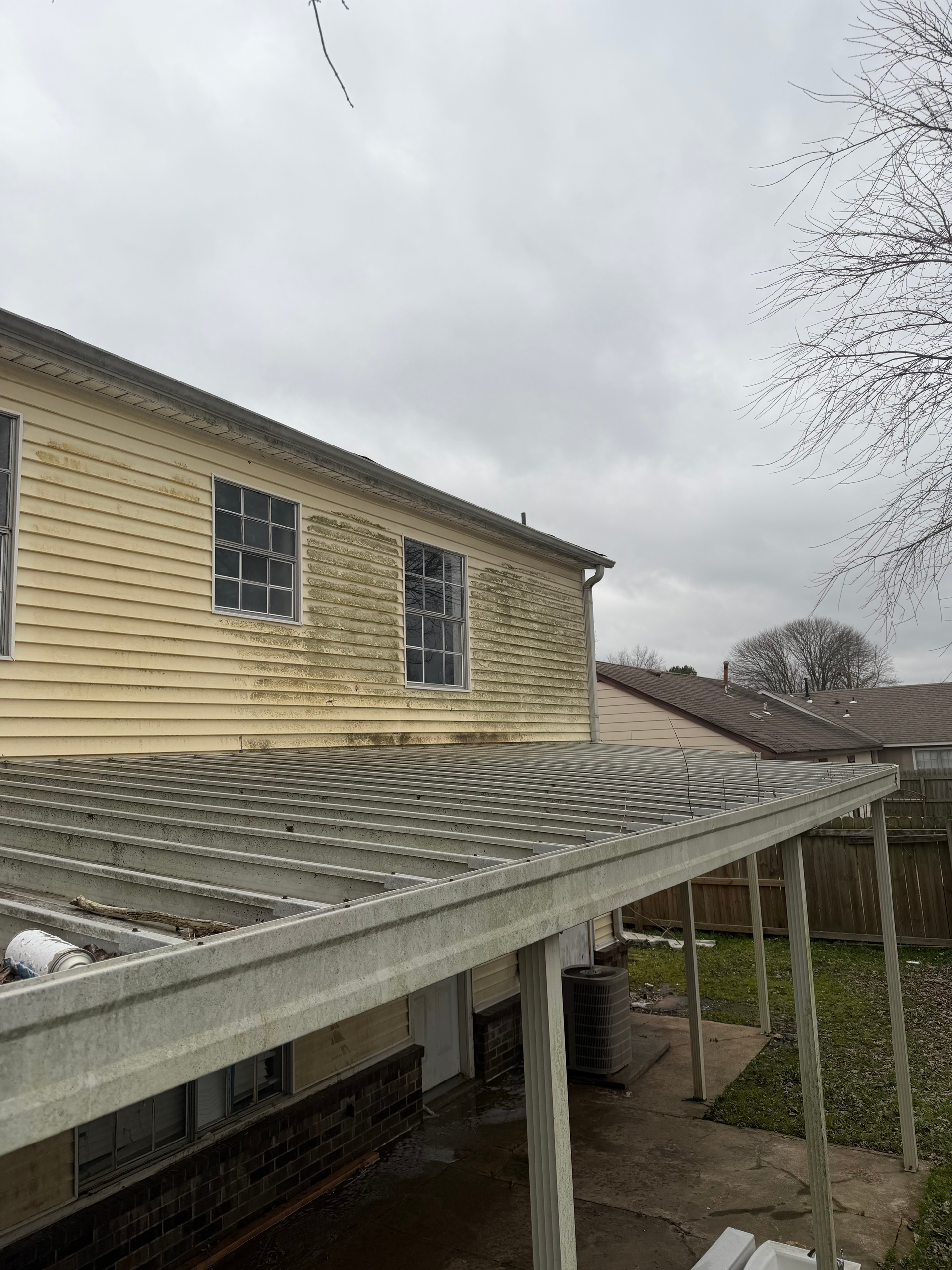 A light yellow two-story house with siding and a gray corrugated patio cover under a cloudy sky.