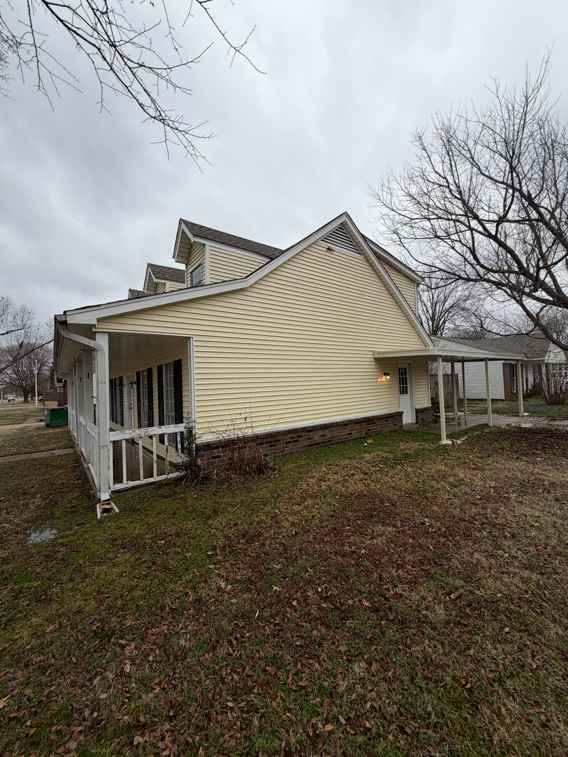 A side view of a light yellow, two-story house with a porch and stone siding base on a grassy, leaf-covered lawn.