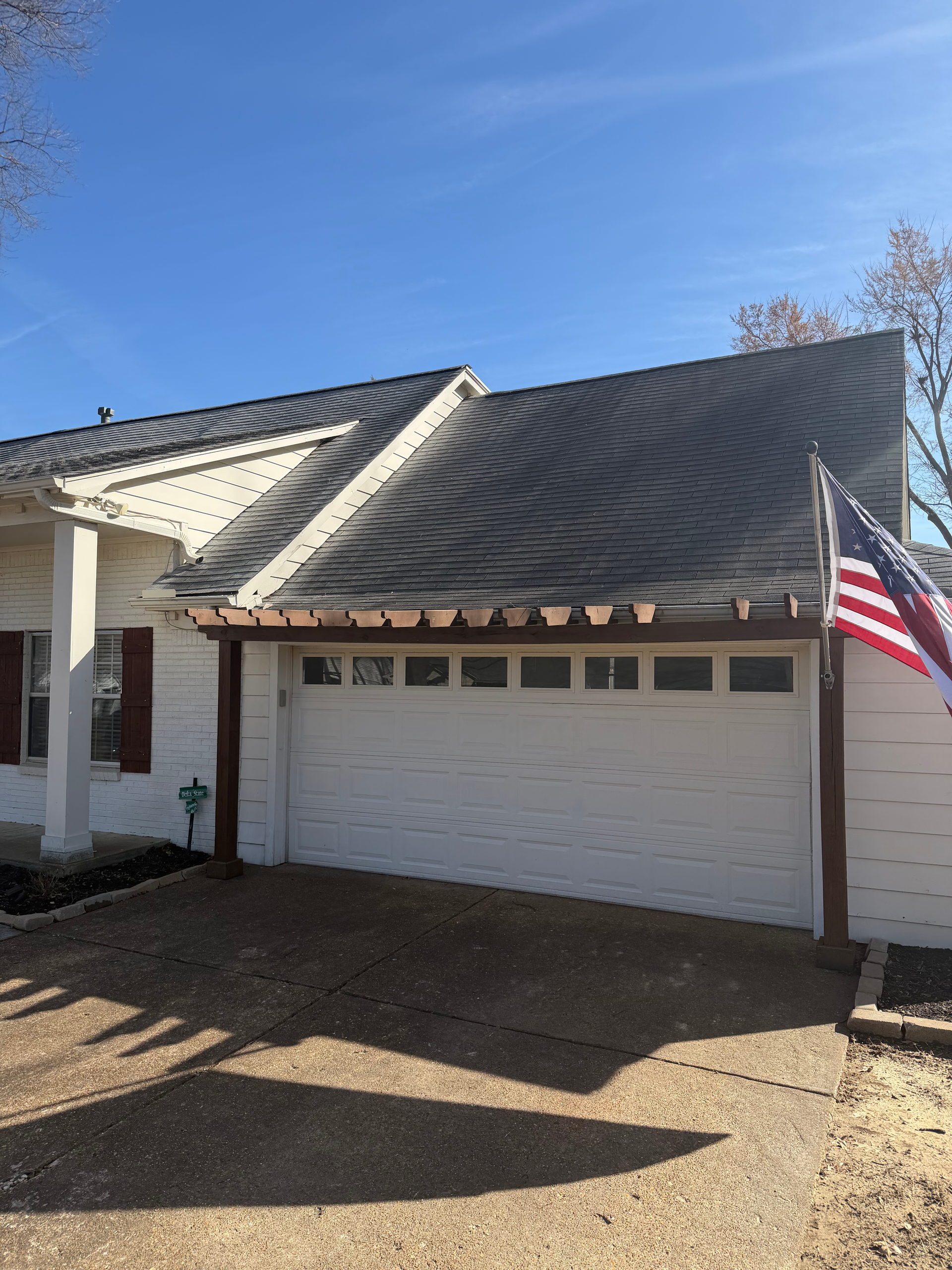 A residential white house with a garage, a shingled roof with dark moss growth, and an American flag attached to a beam.
