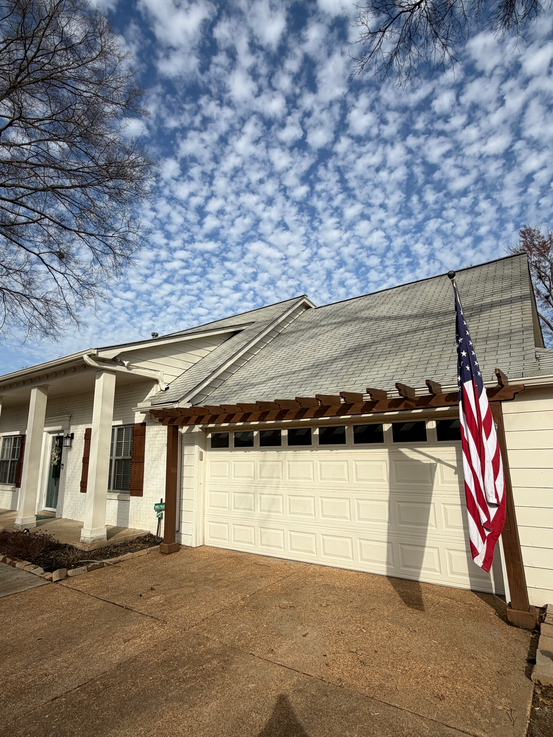 An off-white house with a patterned garage door, wooden pergola, and American flag under a bright, cloudy blue sky.