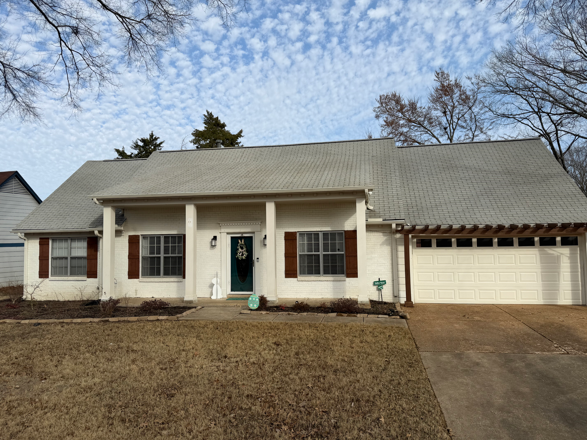 A one-story white brick house with a dark shingled roof, a covered porch, and an attached two-car garage under a trellis.