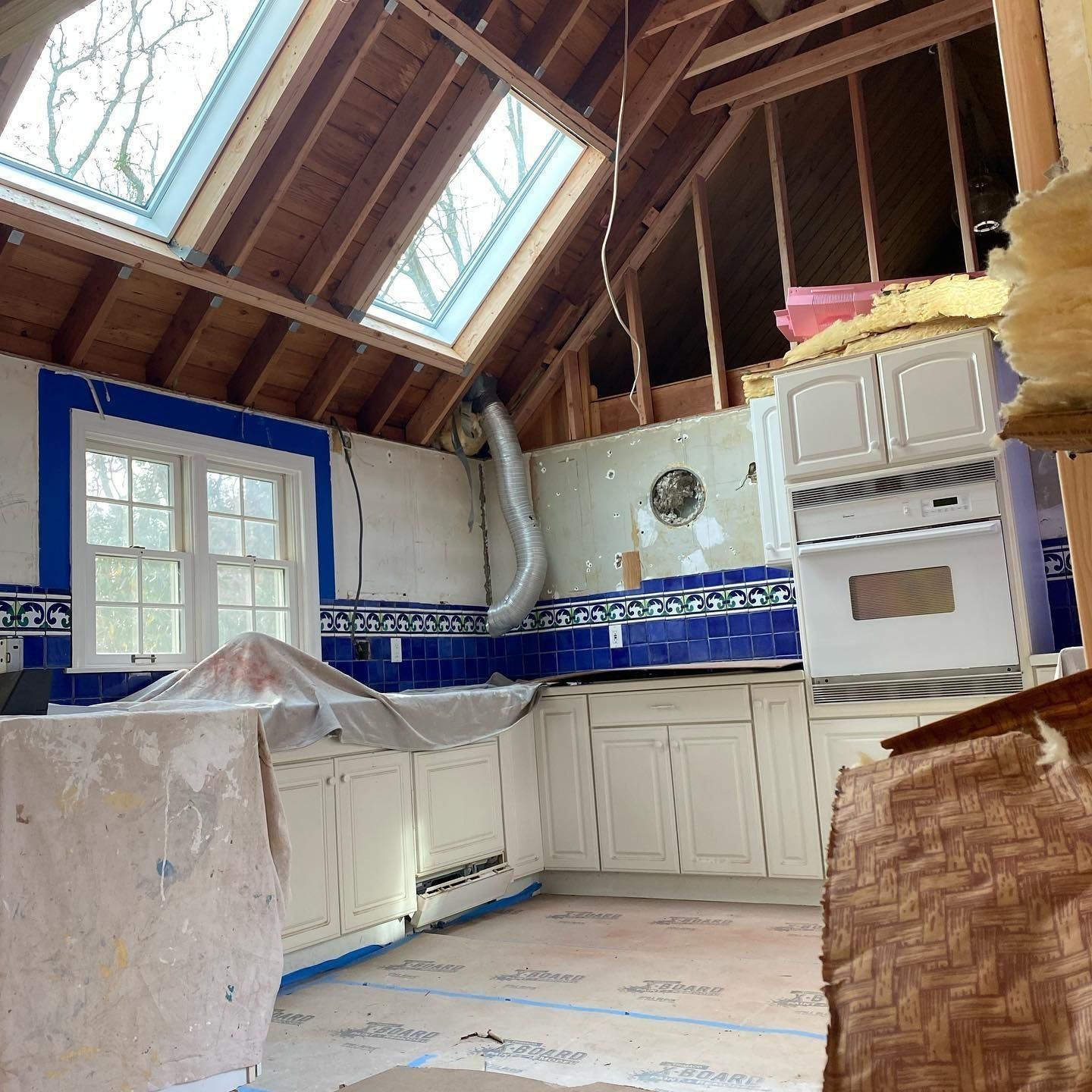 Kitchen under renovation, white cabinets, blue tile backsplash, exposed ceiling beams, skylights, tarp-covered counter.