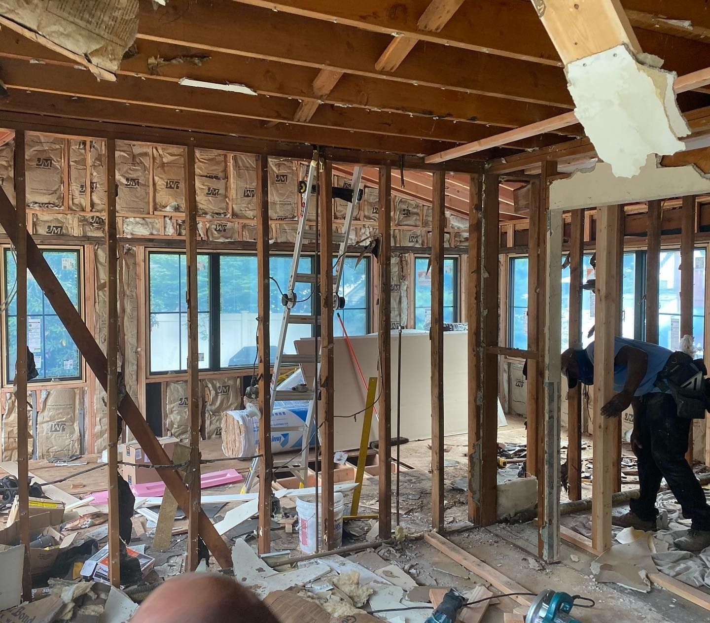 Interior of a building under renovation, exposed wooden framing, insulation, debris on floor, and two workers present.