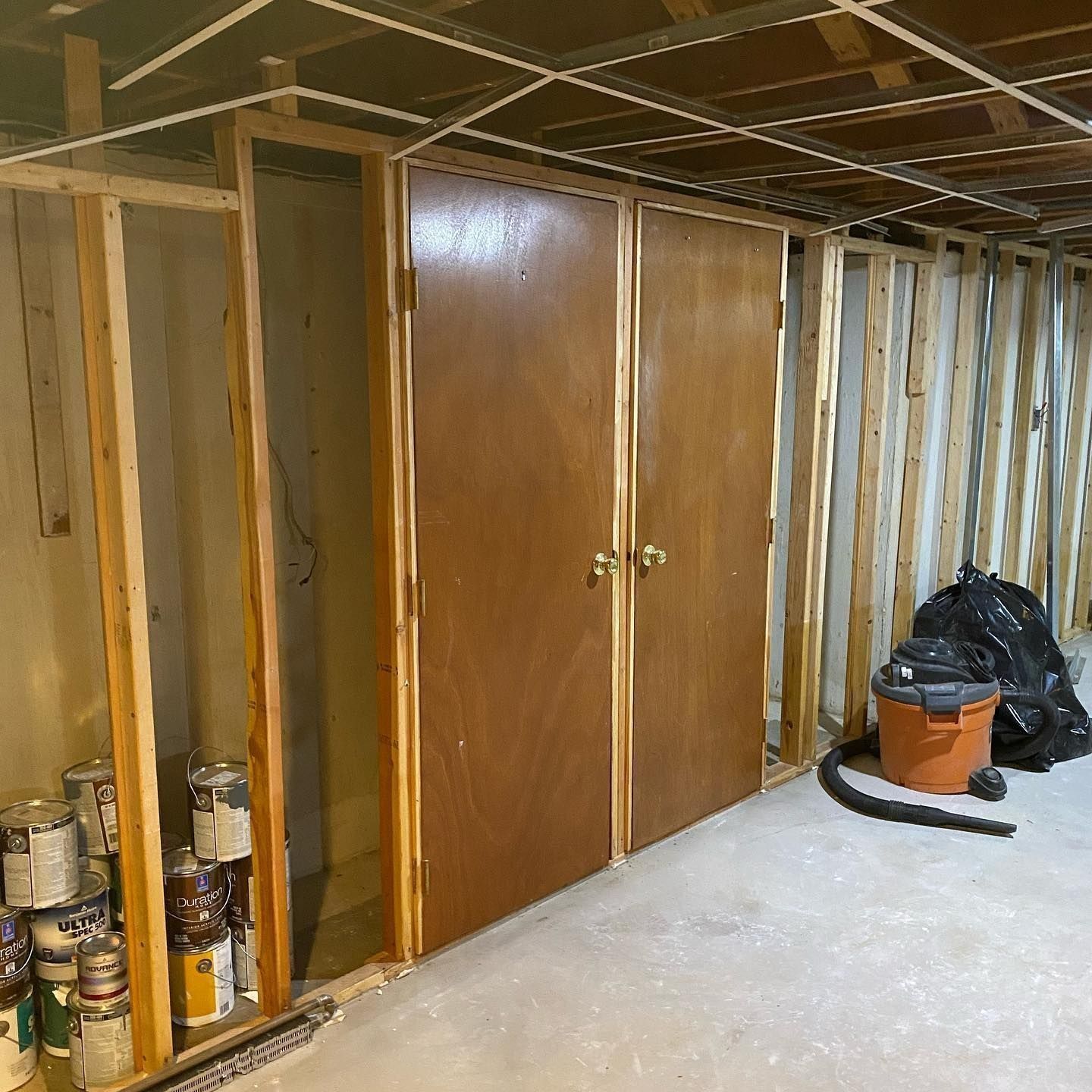 Basement with wooden framing, closet doors, paint cans, and a vacuum.