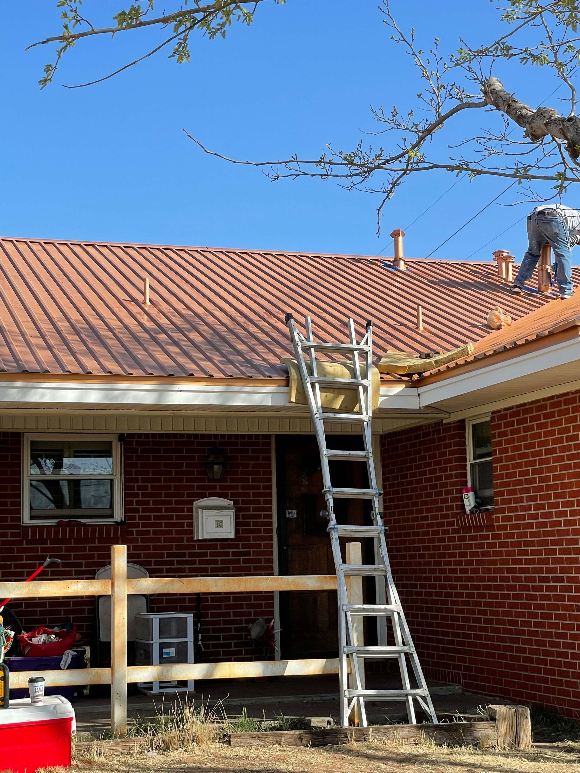 A person on a red tile roof, working with a ladder against the house. Brick building, blue sky.