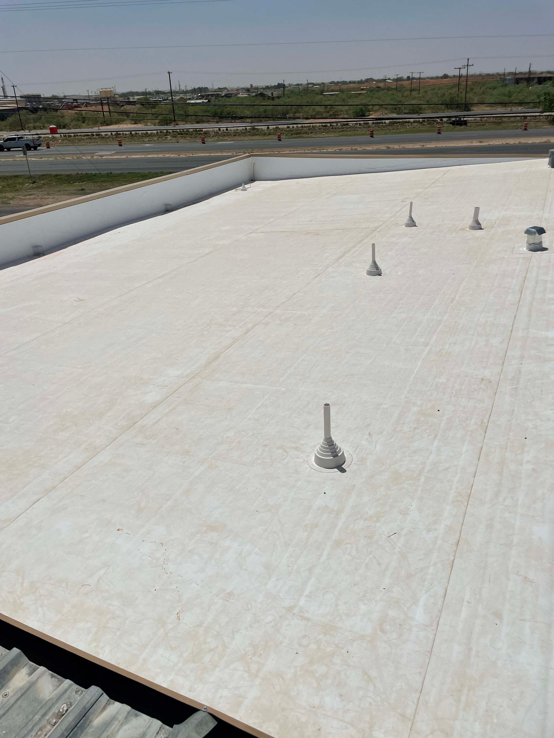 View of a flat, white commercial roof with ventilation pipes, against a background of a cityscape and sky.