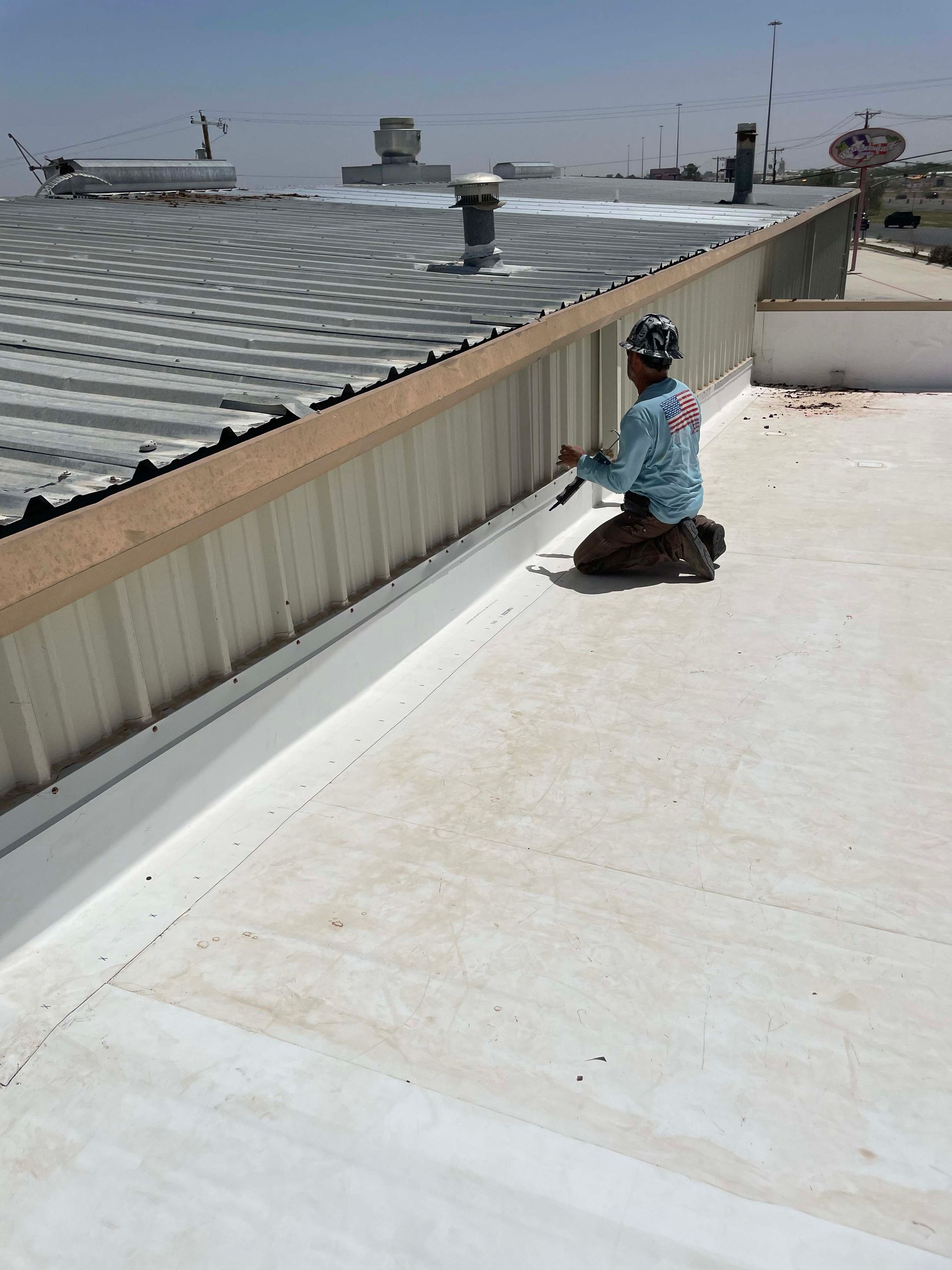 Person applying white coating to a flat roof. Metal trim and buildings in the background. Bright sunlight.