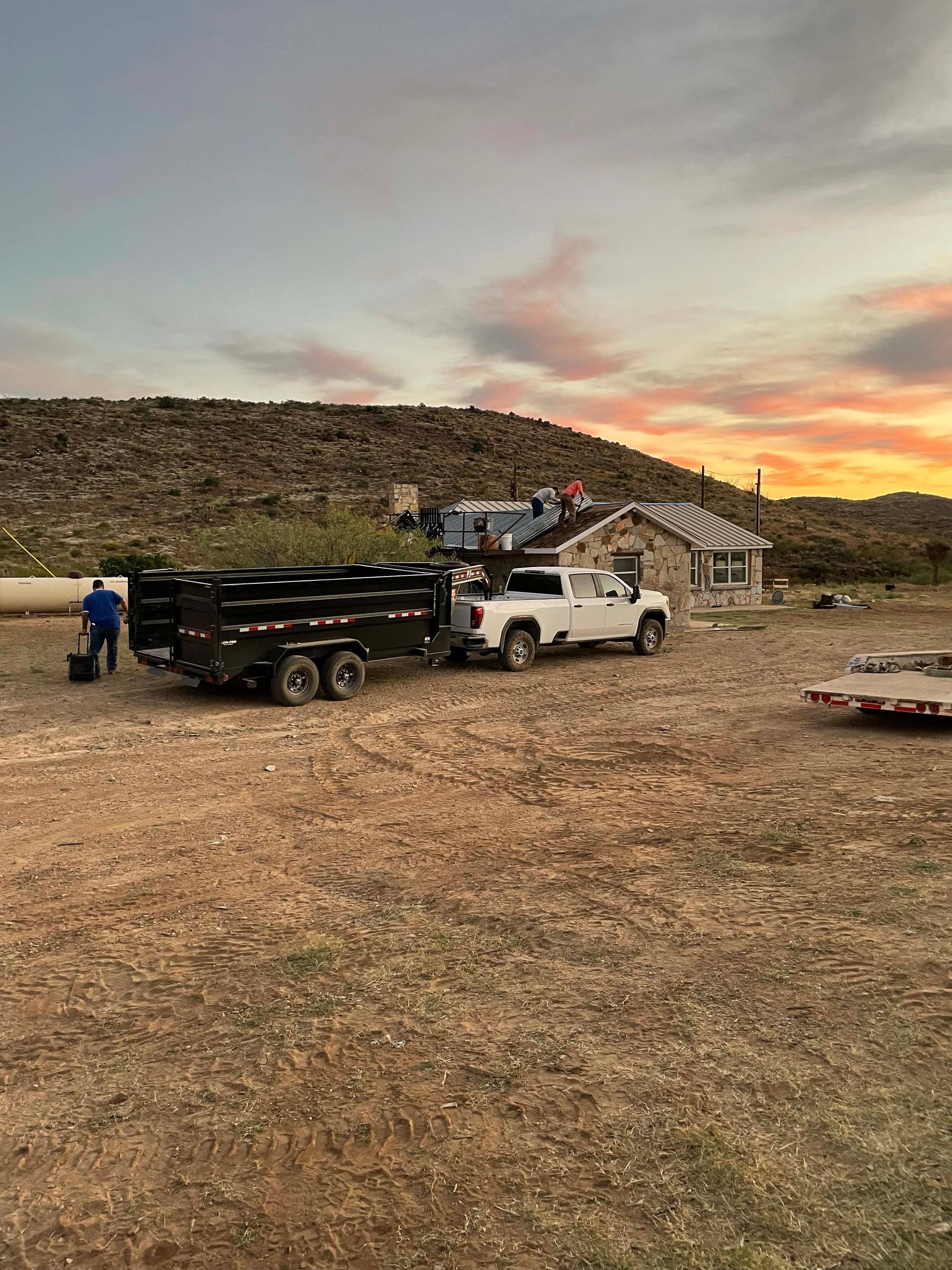White truck with a trailer, parked in front of a building. Two people are nearby, with a sunset in the background.