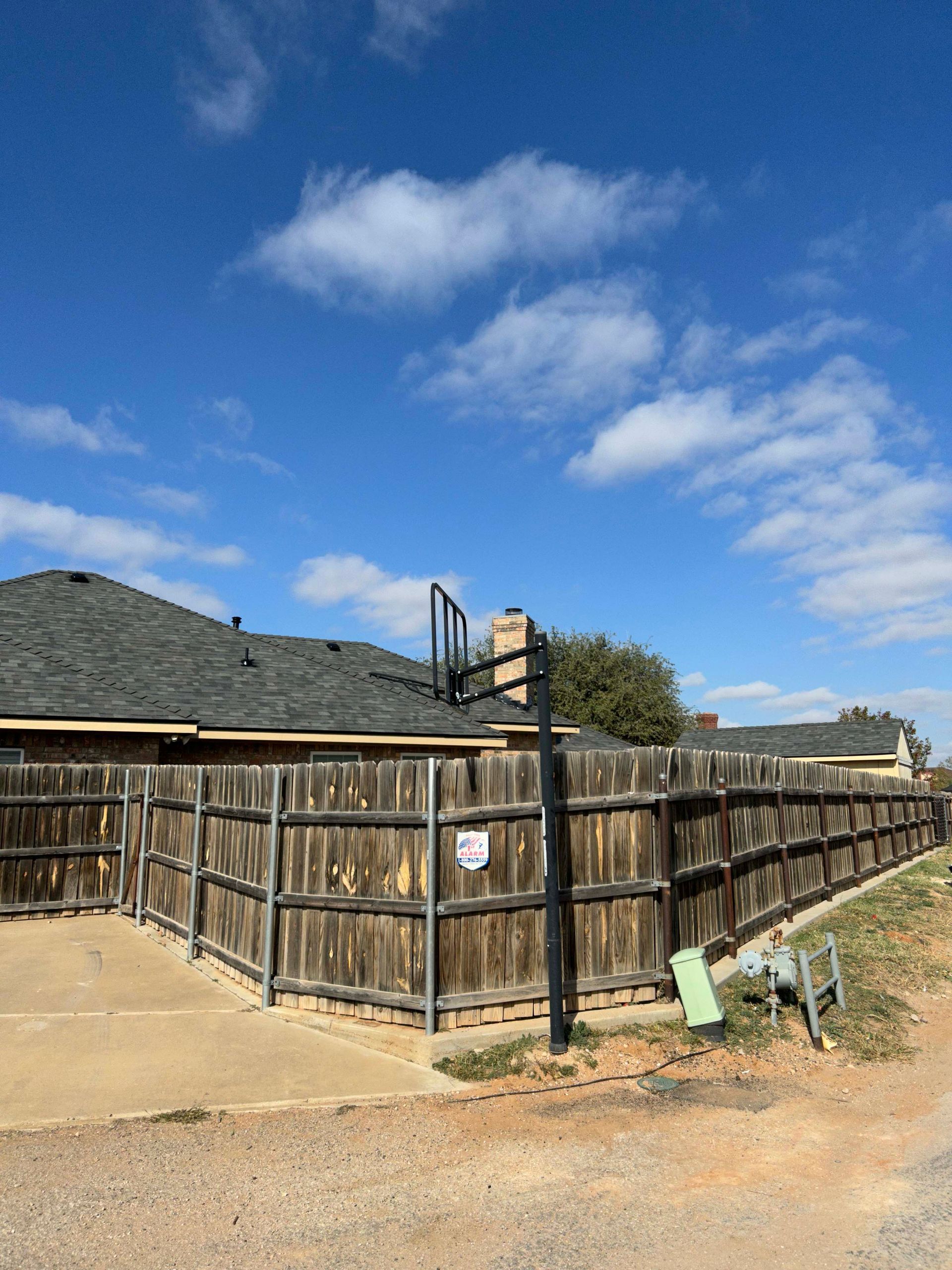 Wooden fence surrounds a house with a basketball hoop on a sunny day.