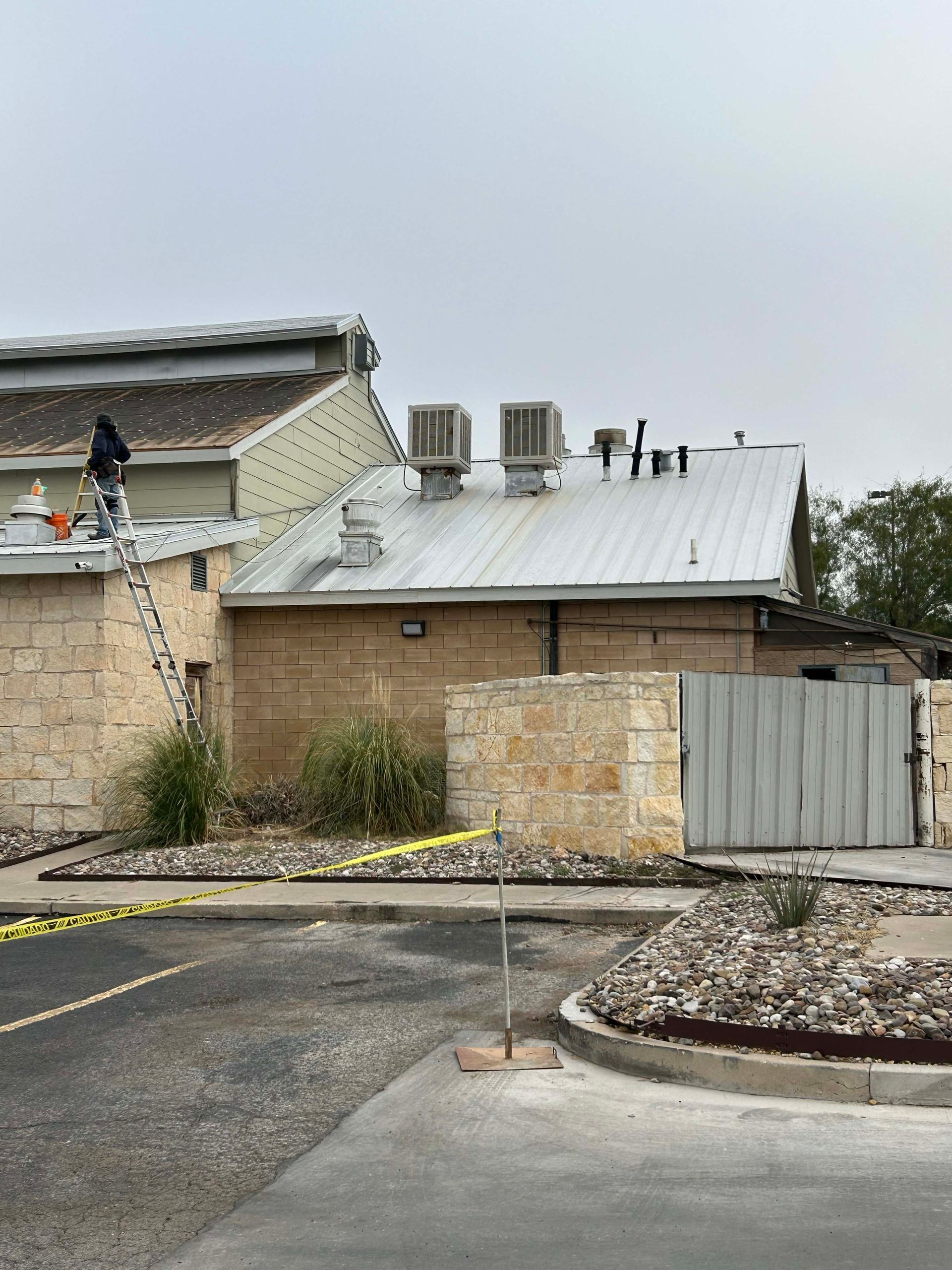 Person on a ladder repairs a metal roof. Beige stone building, cloudy sky, caution tape.