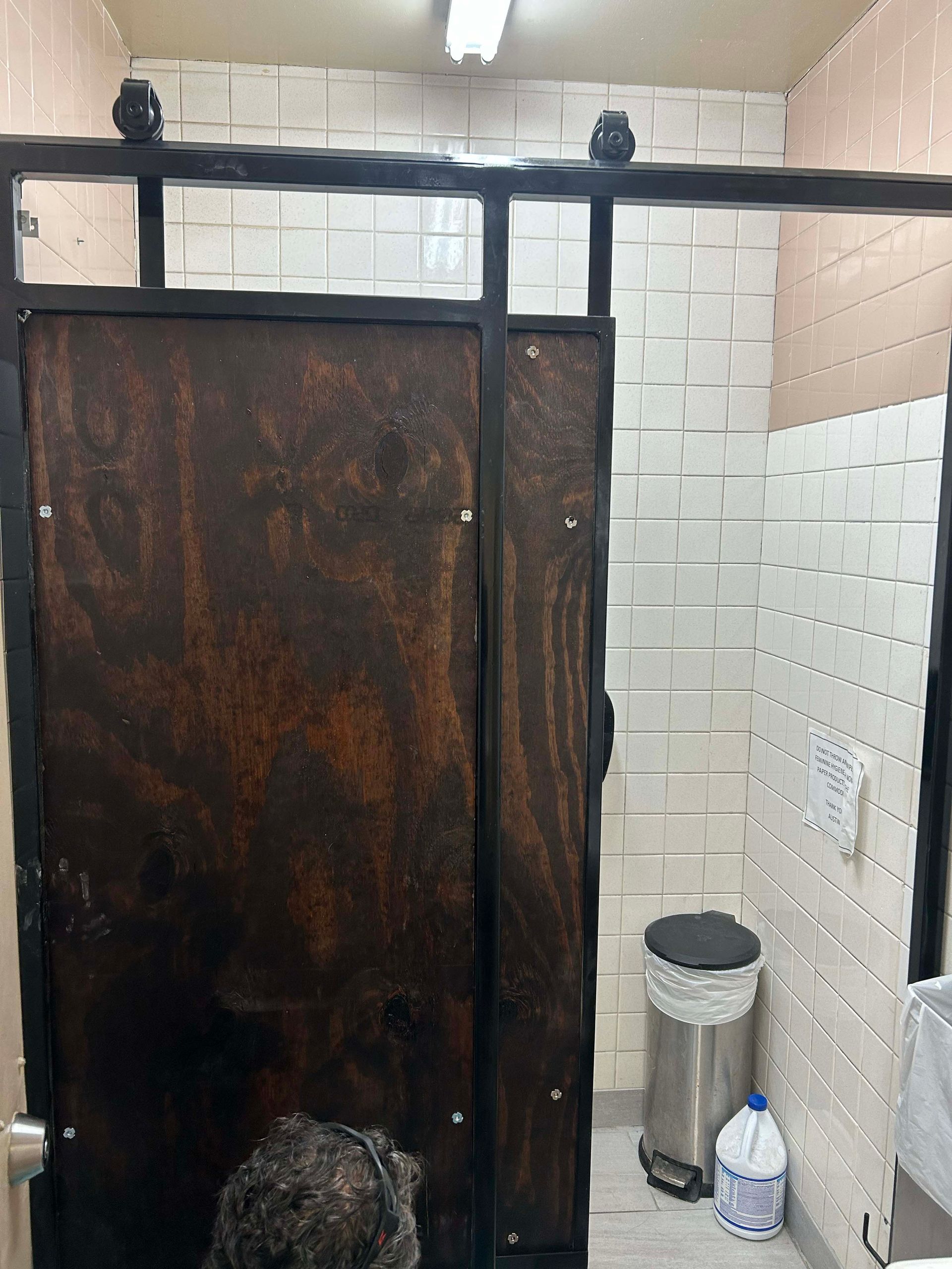 Bathroom stall with dark wood door and black metal frame, white tile walls.
