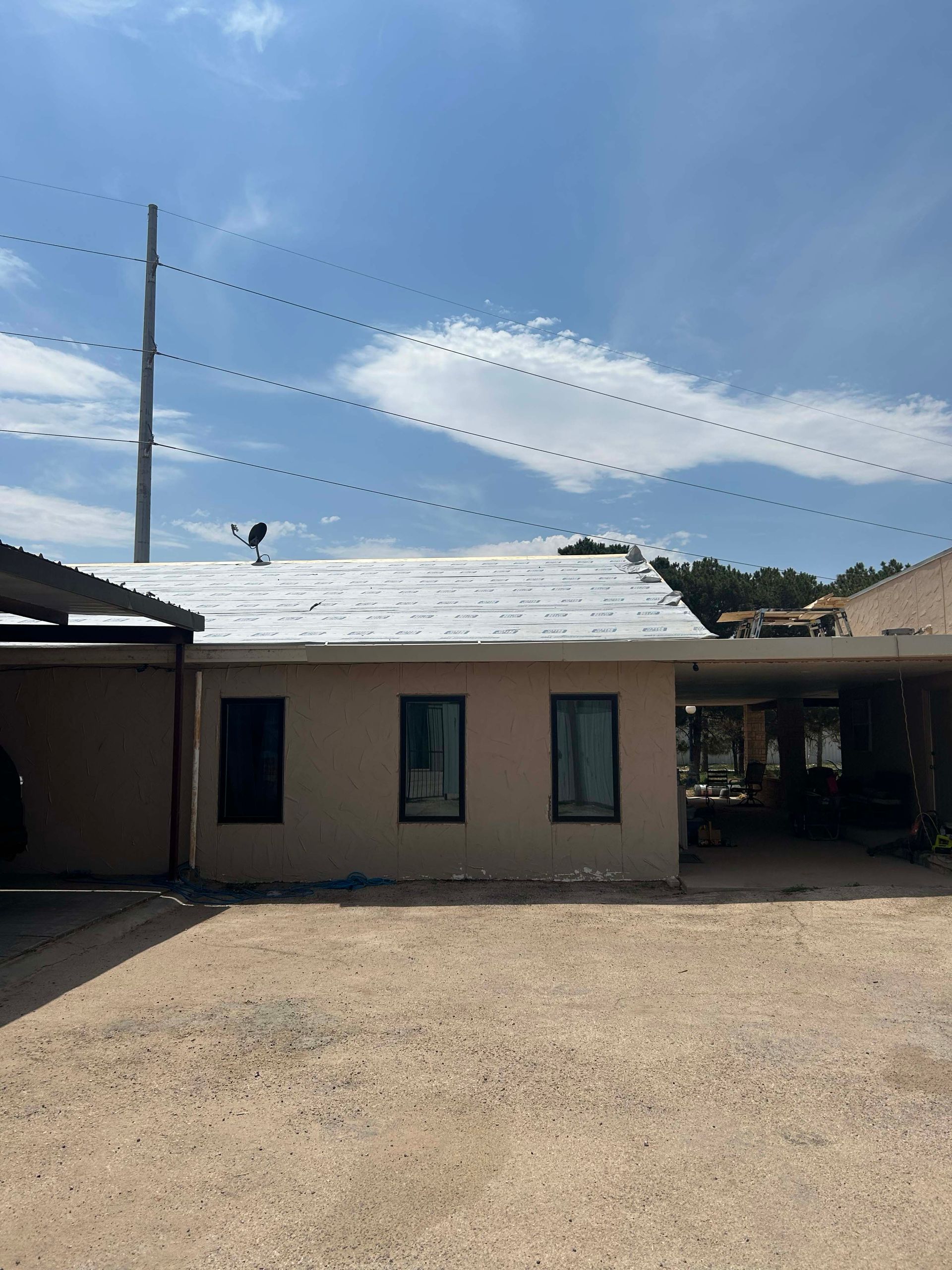 Beige building with three windows, carport, and power lines against a blue sky.