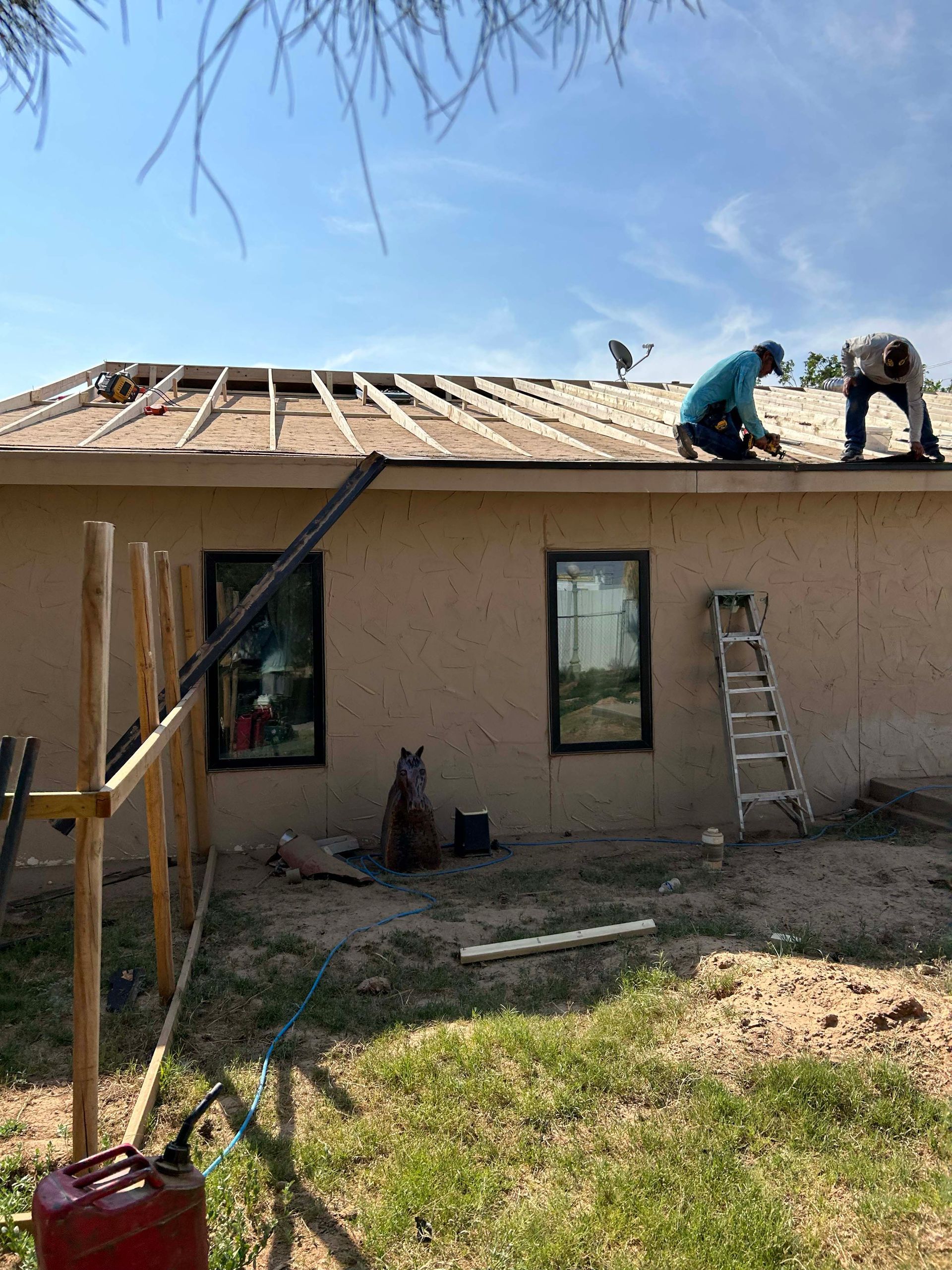 Two people working on a roof, near a tan building with dark window frames. A ladder is present.