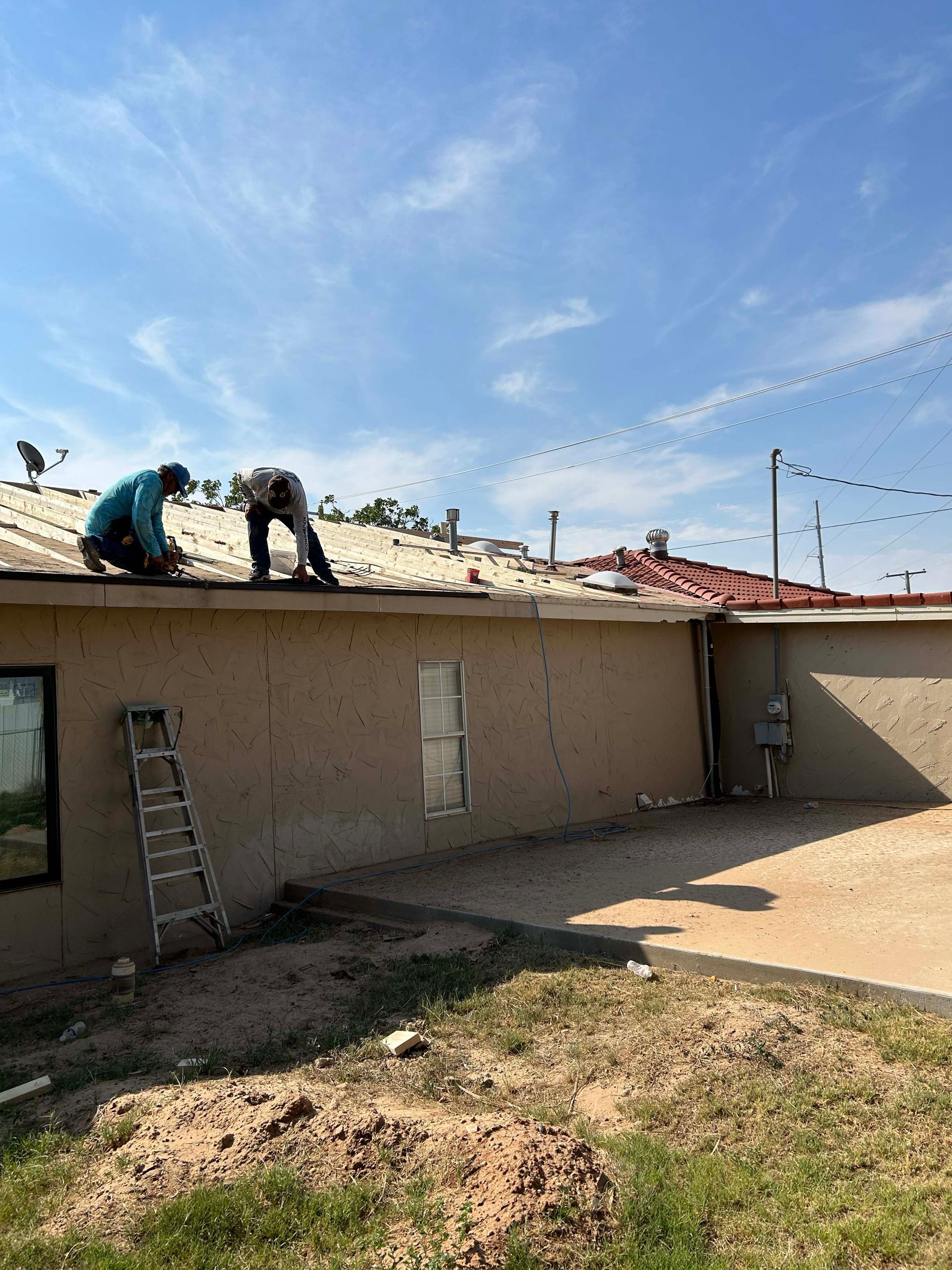Two people on a roof working on shingles under a blue sky, with a ladder and the side of a building visible.