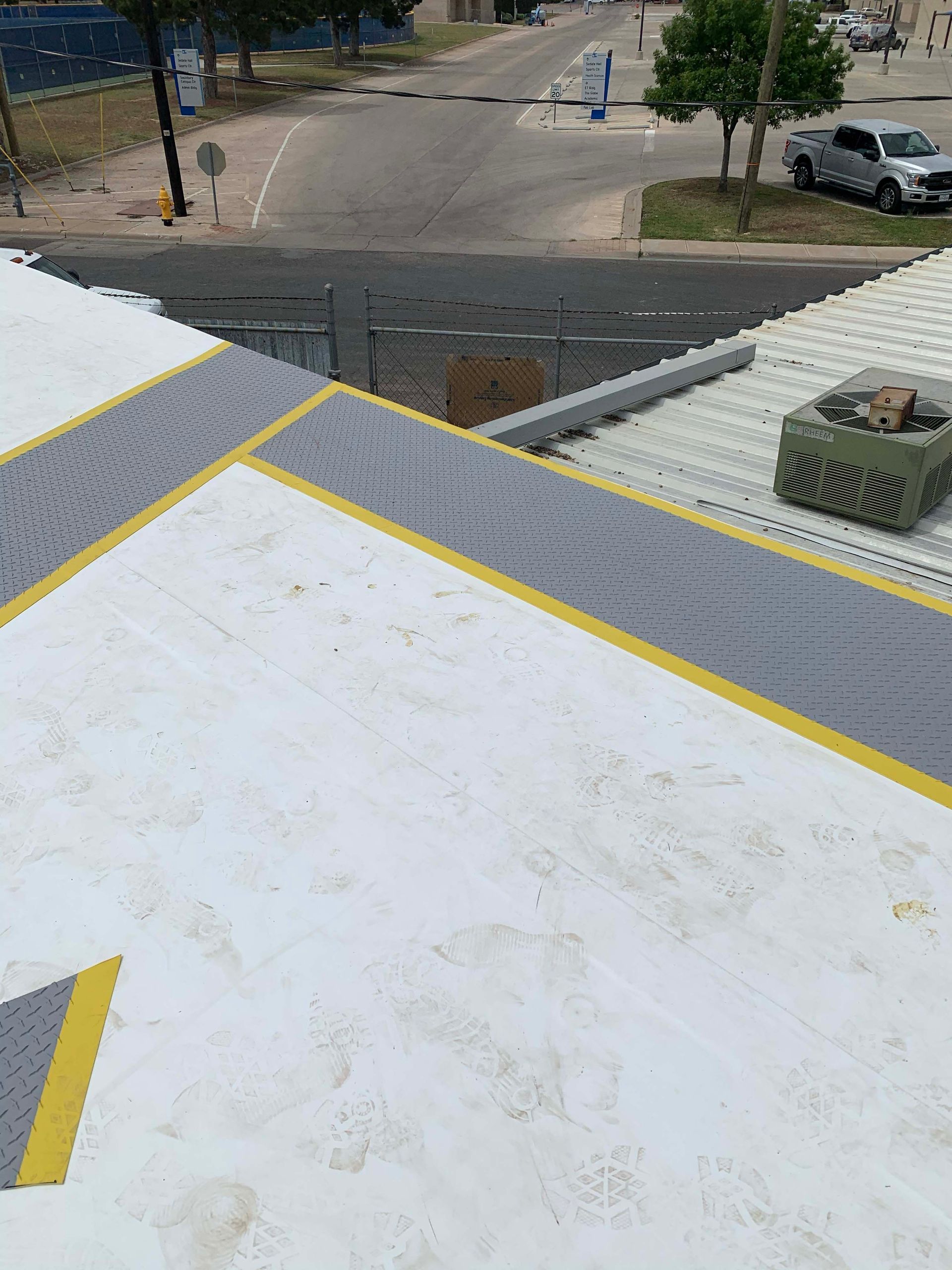 White commercial roof with gray and yellow walkways and an HVAC unit. City street in background.