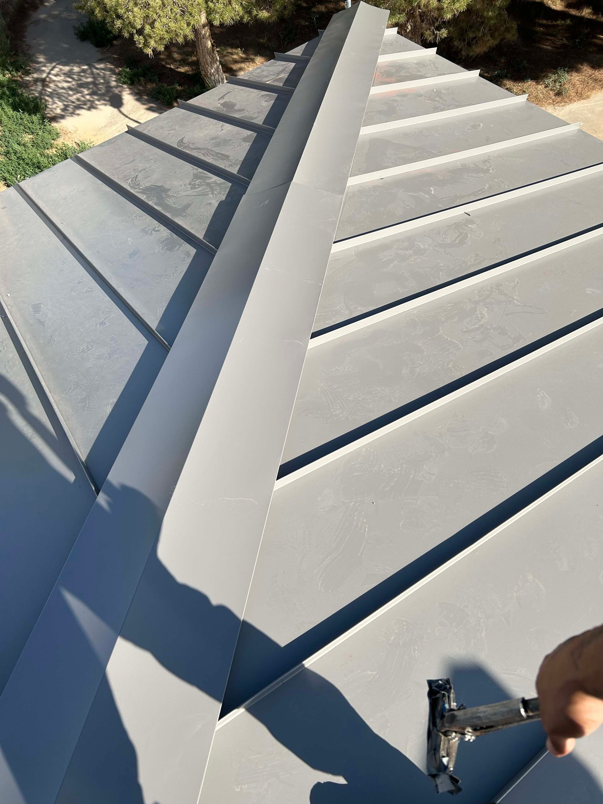 Gray metal roof being installed, close-up. A hand holds a tool near the ridge.