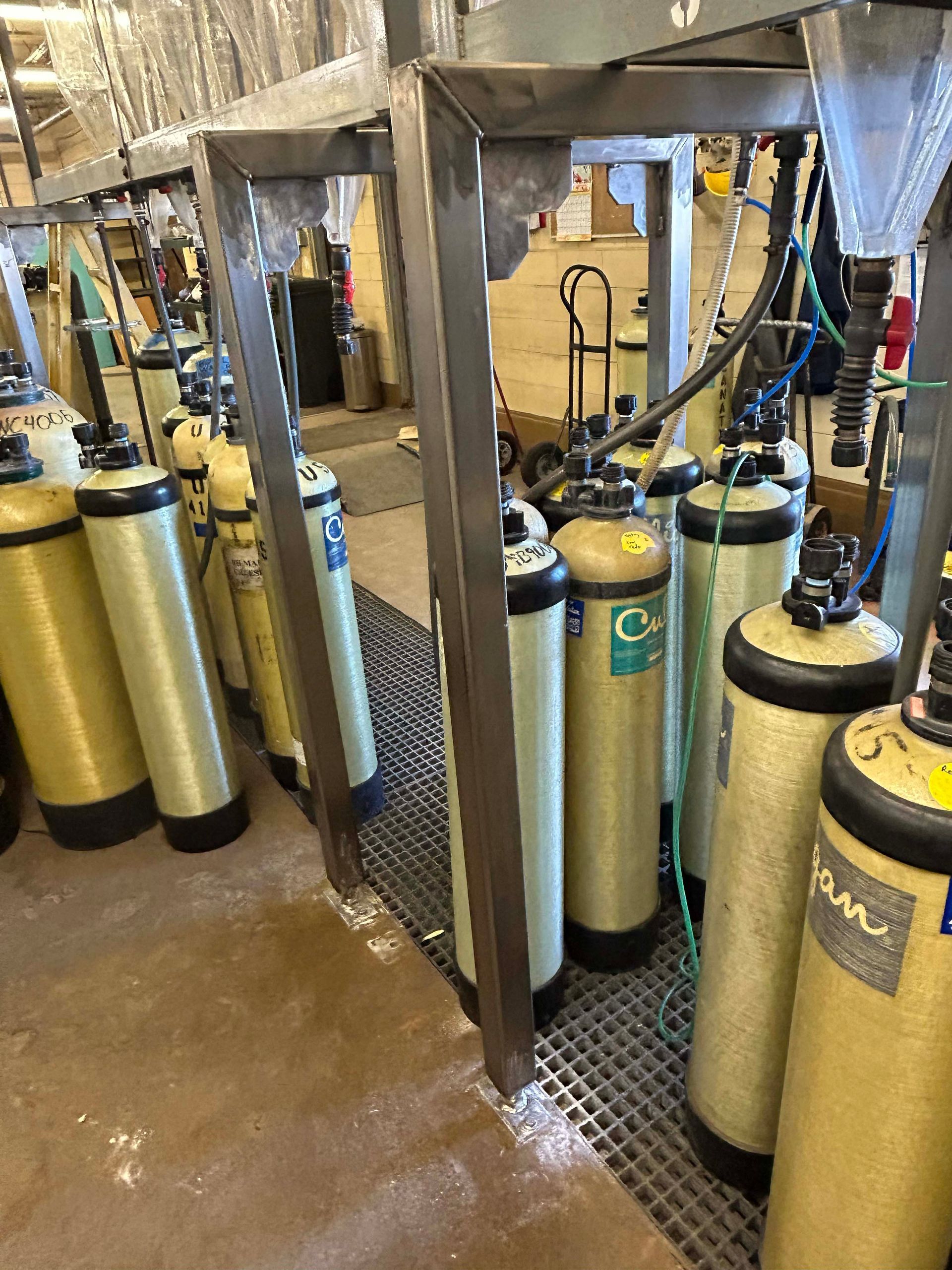 Tanks lined up within metal frames, likely in an industrial setting. Some tanks are yellow, green, and blue.