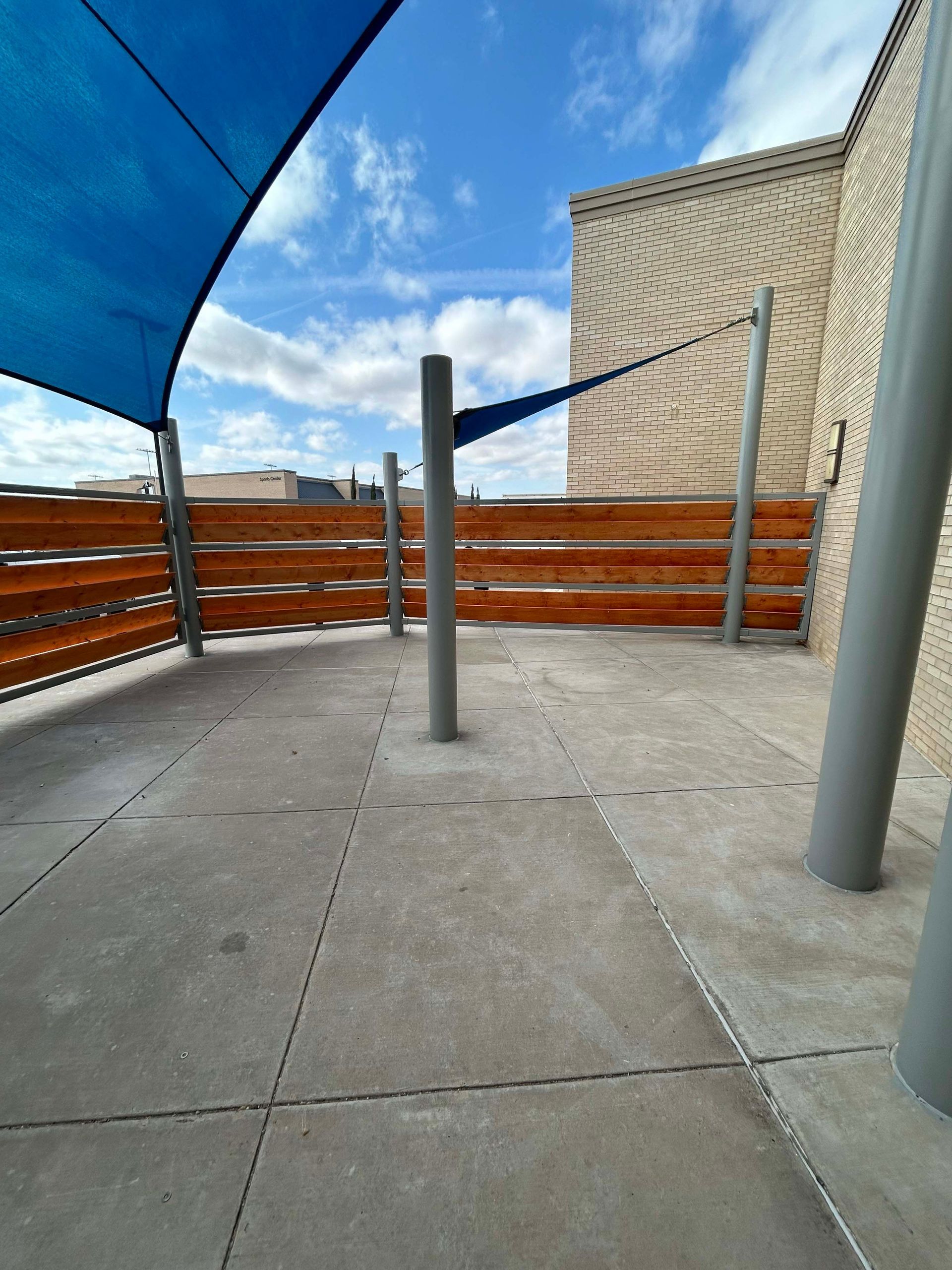 Outdoor patio with blue shade canopy, wood fence, gray pillars, and a cloudy sky.