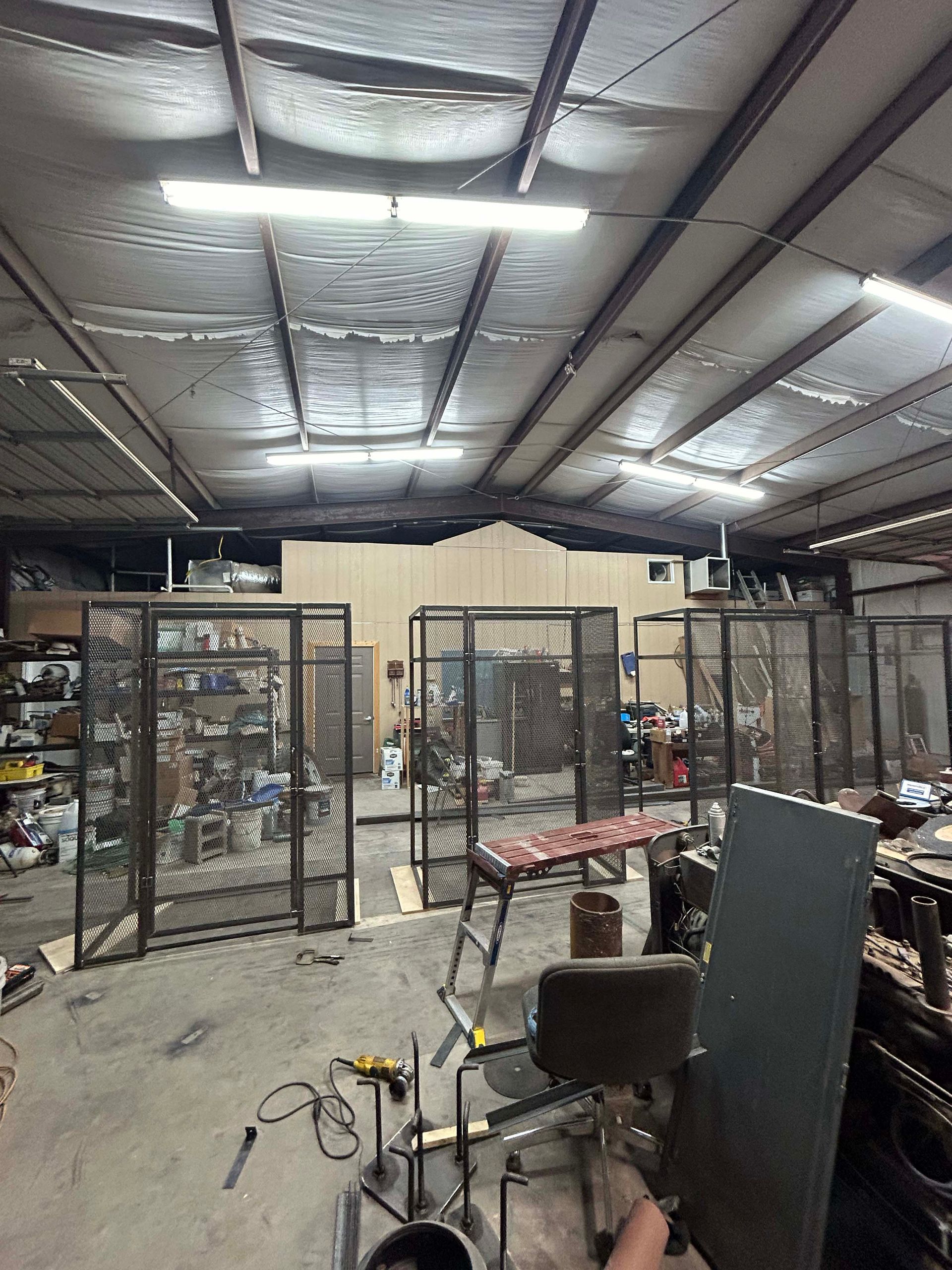Metal fabrication workshop interior with several caged storage units, fluorescent lighting, and tools.