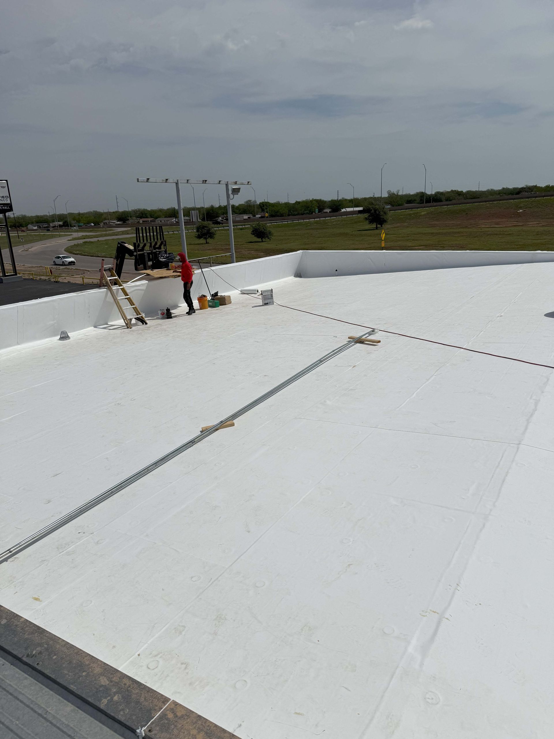 Workers on a white commercial roof, applying sealant. Safety equipment in place, clear sky.