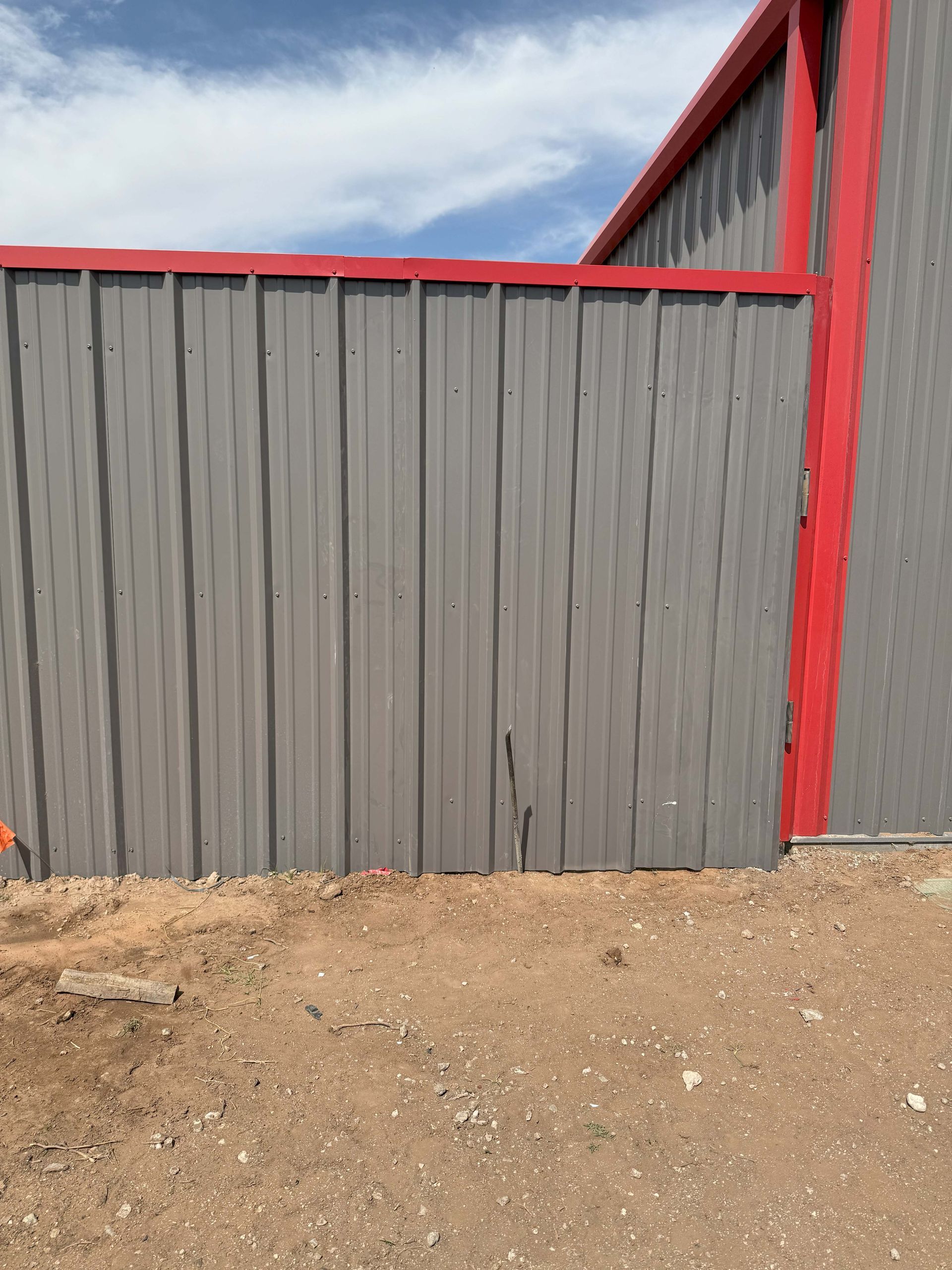 Gray metal building with red trim, set on dirt ground with a blue sky.