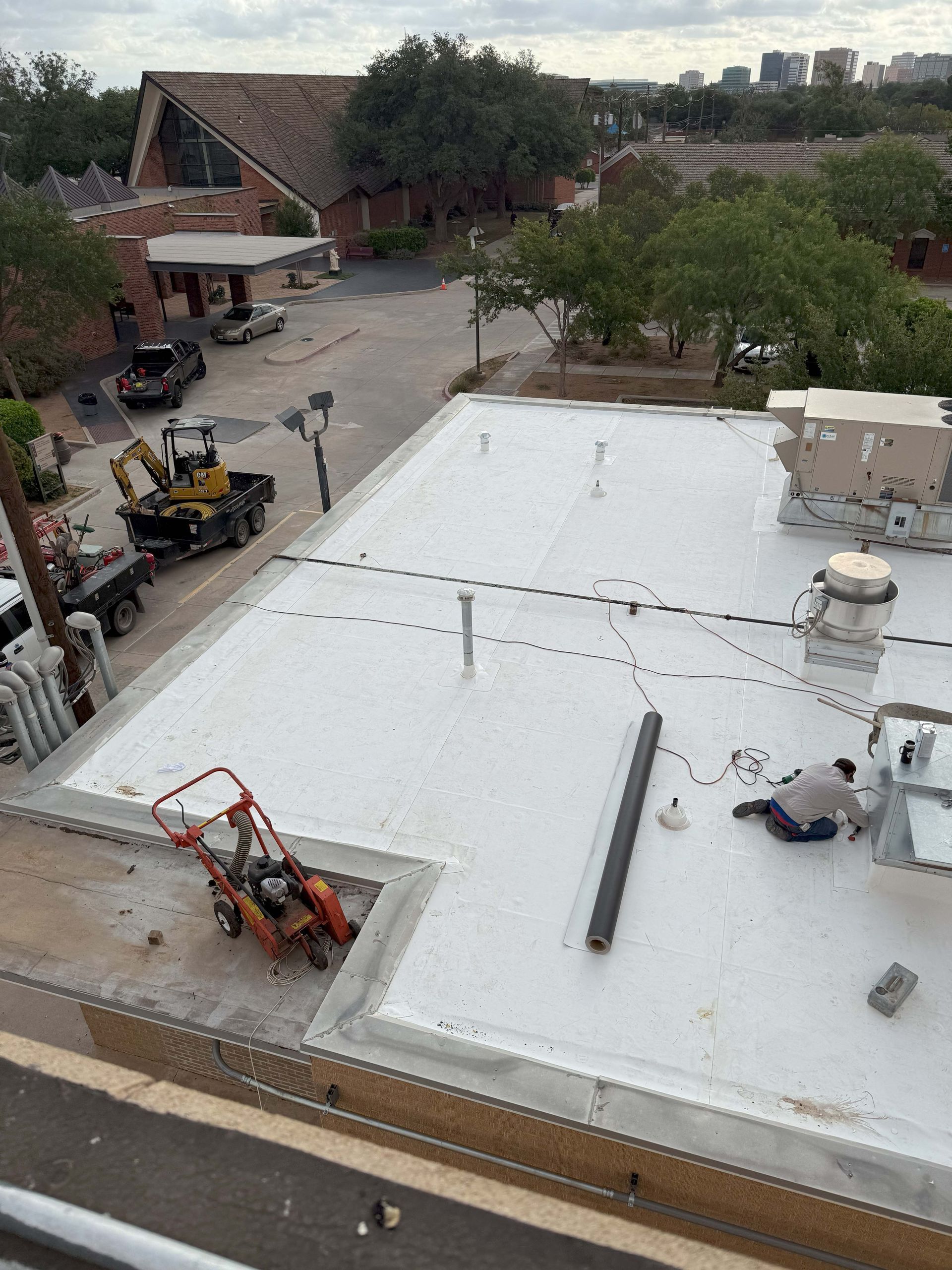Rooftop construction: workers, machinery, white surface, buildings, trees, cloudy sky.
