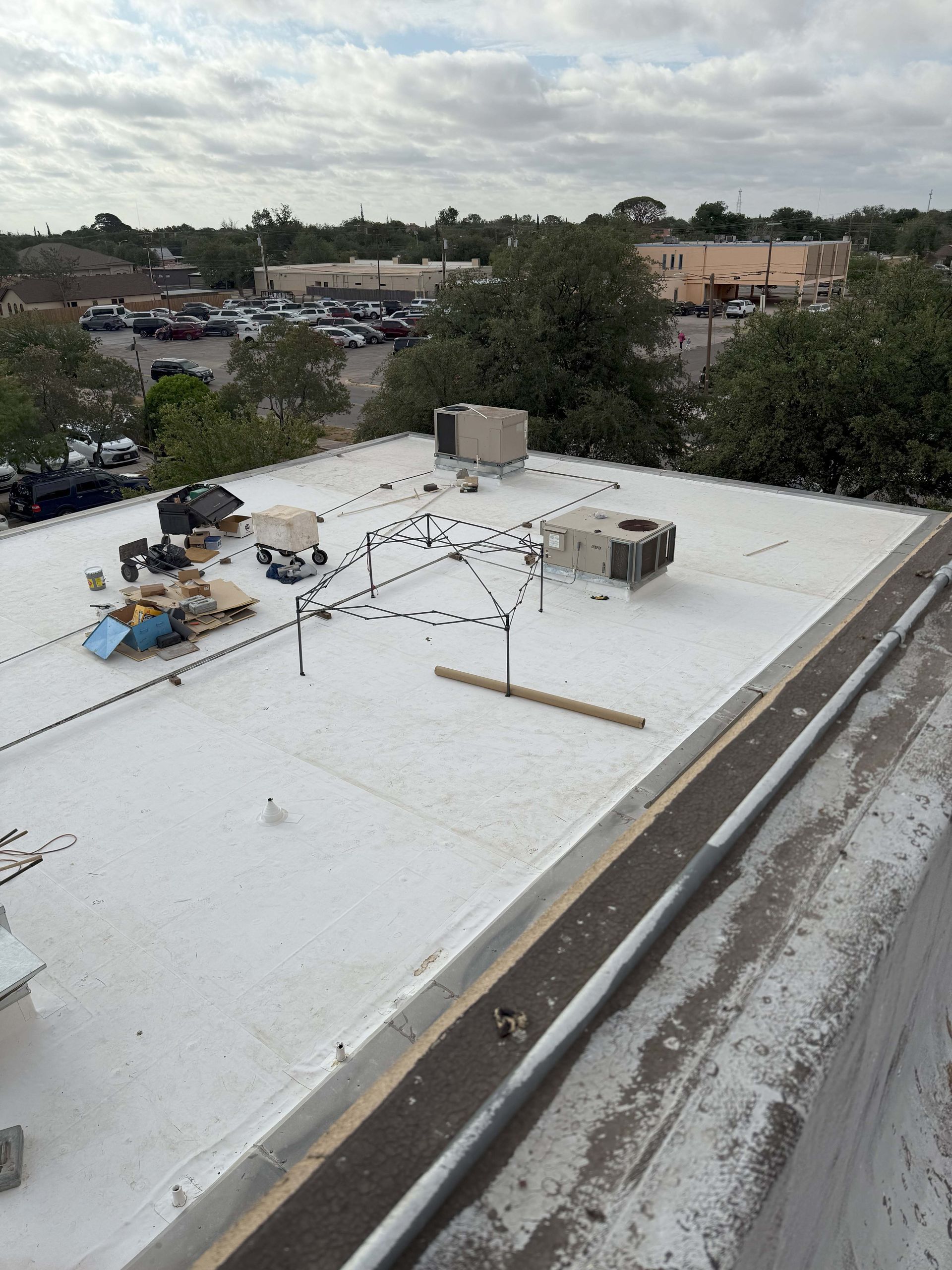 White roof of a building with equipment and surrounding parking lot, trees, and sky.