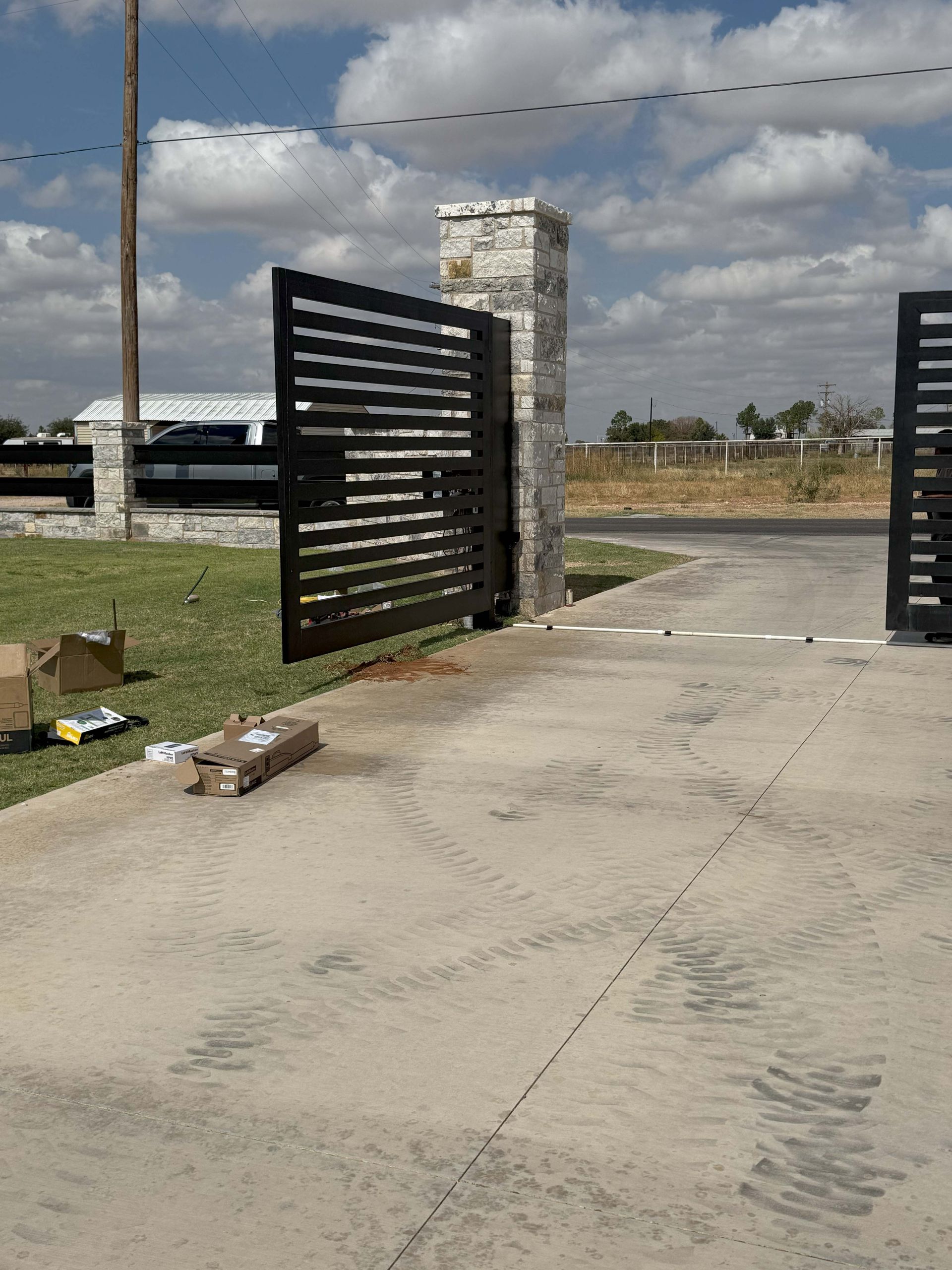 Black metal driveway gate, open, with stone pillar on a sunny day.