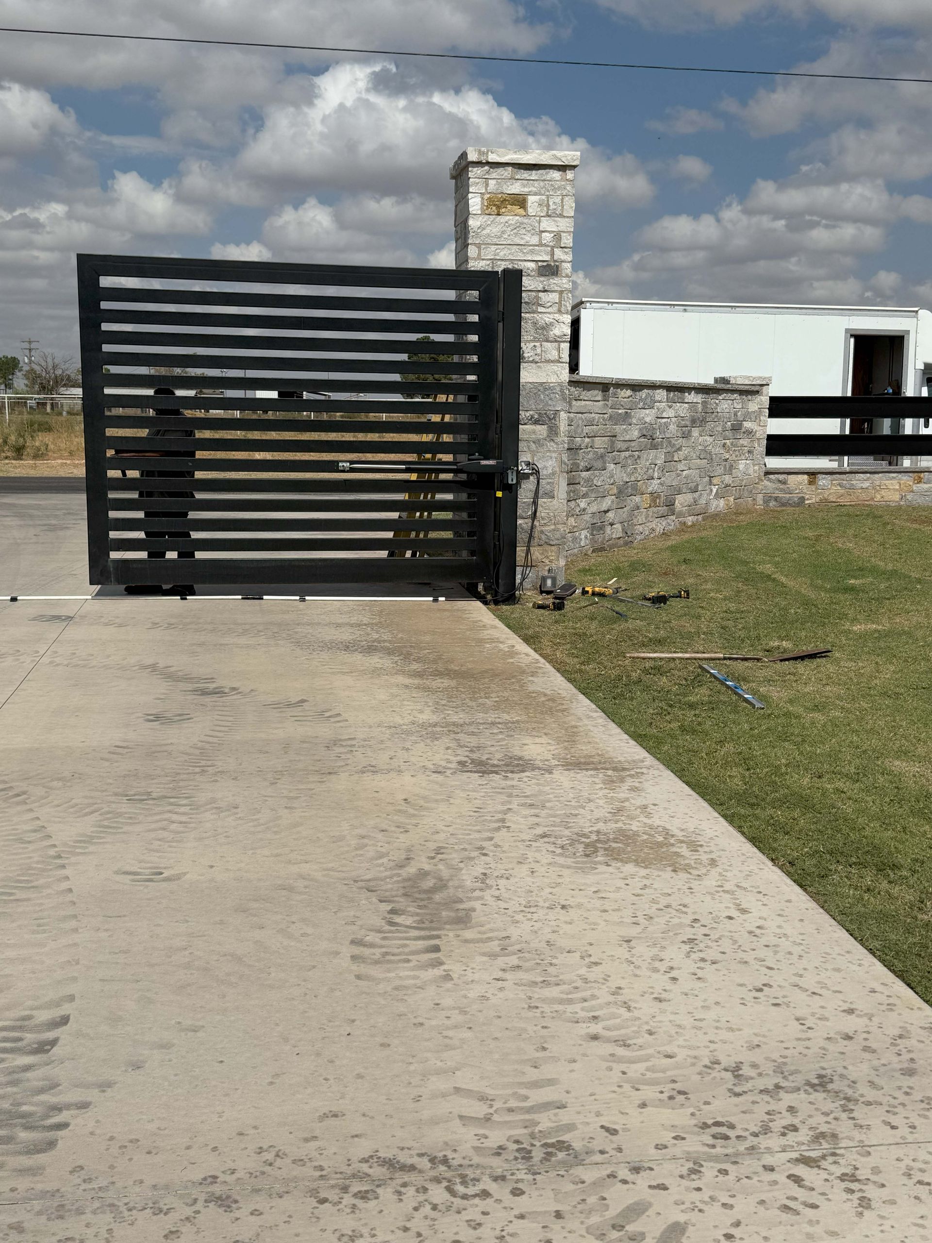 Black metal gate and stone wall on concrete driveway. Buildings and green lawn in background.