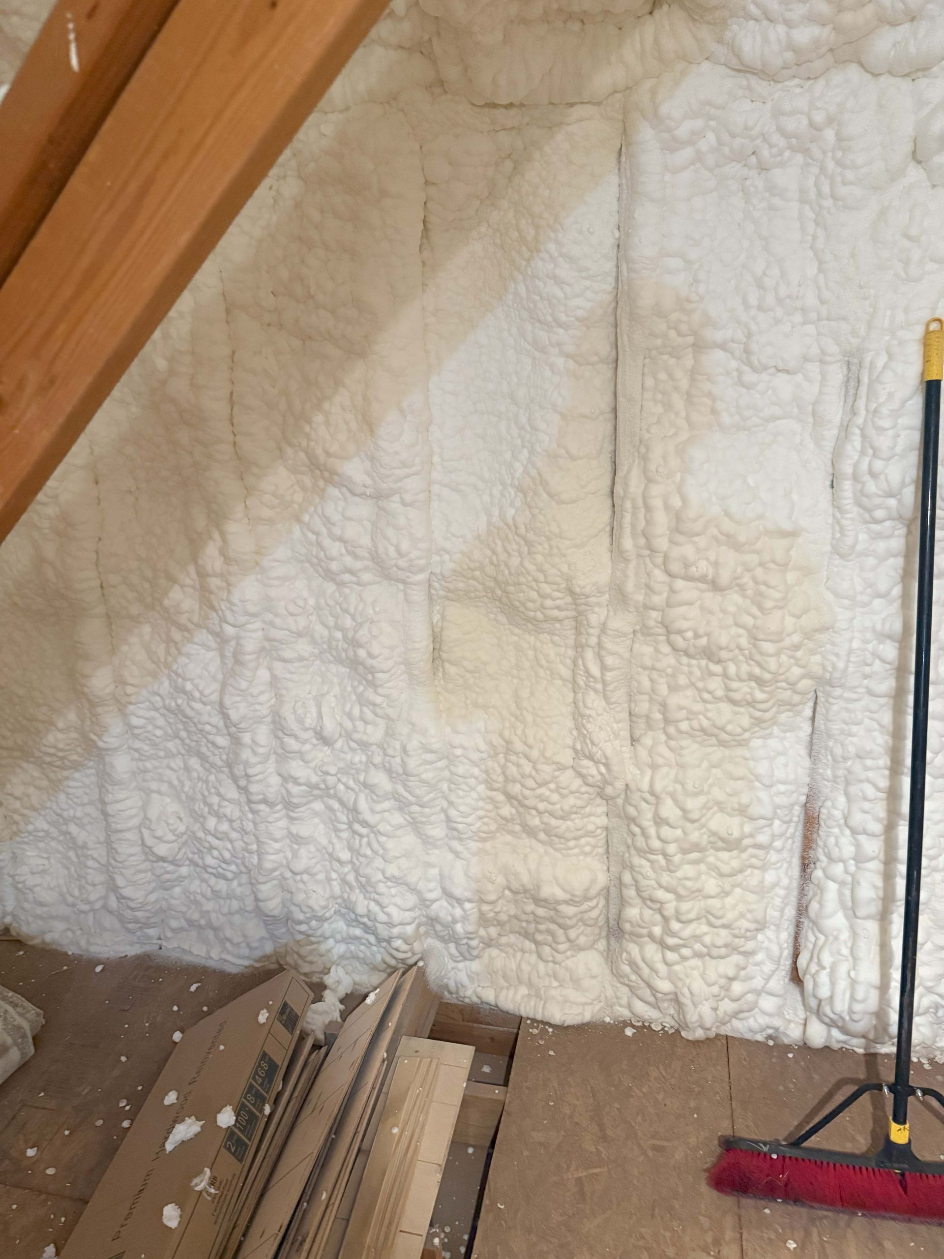 Attic wall insulated with white spray foam; wooden beams, broom, and cardboard visible.