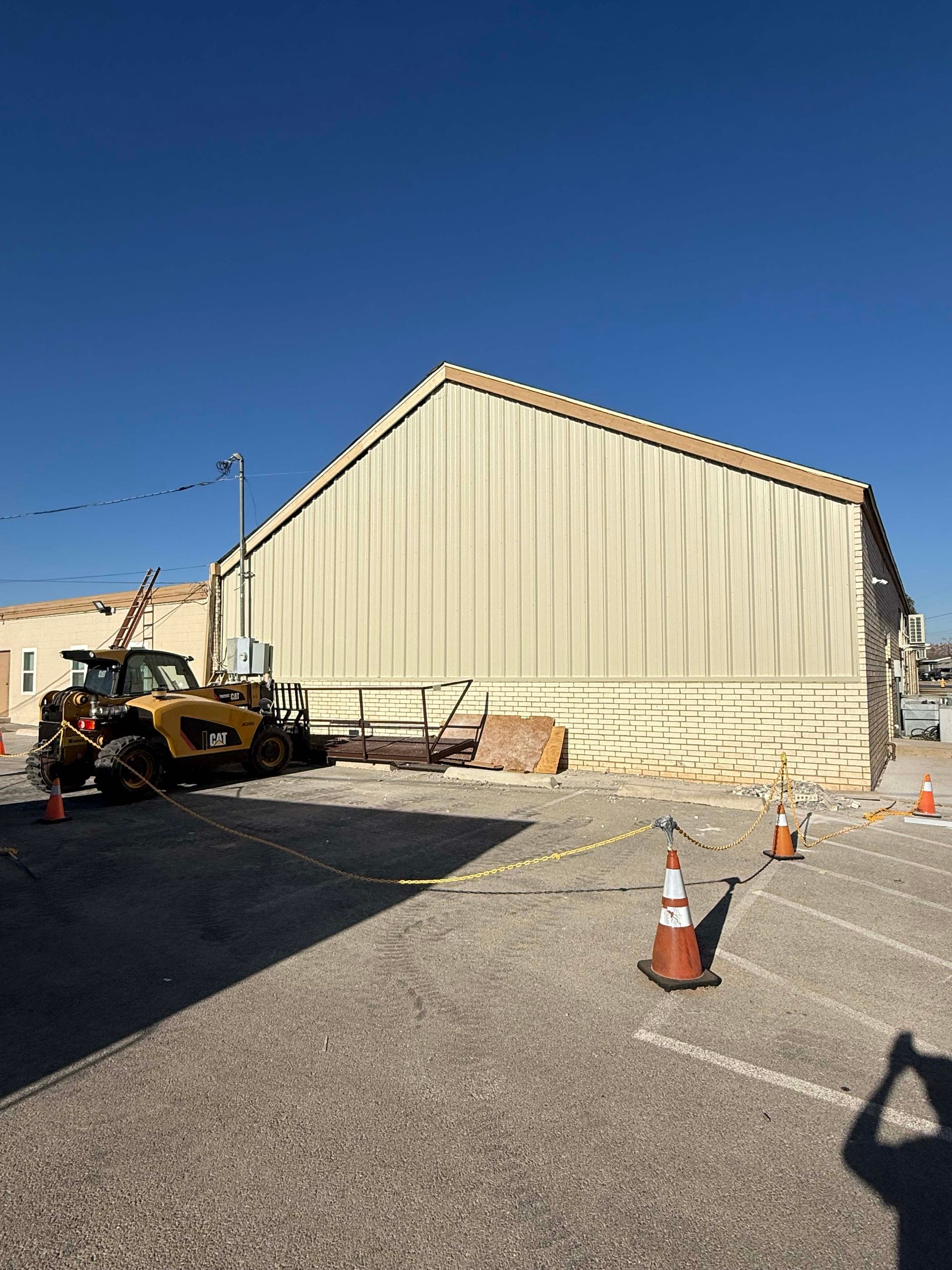 Yellow construction vehicle near a beige metal building with safety cones in a parking lot under a blue sky.