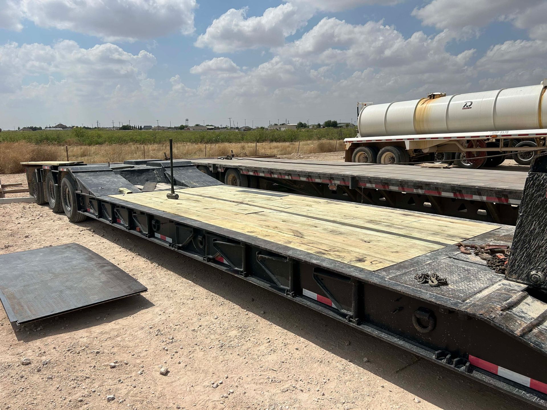 A black flatbed trailer with a light-colored wooden deck in a dusty outdoor setting.