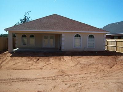 Beige house with arched windows and brown roof, set in a dirt yard with a fence.