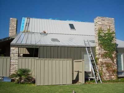 House with metal roof under construction; ladder, stone chimneys, and tan siding.