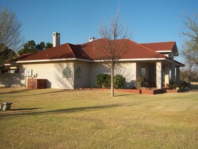 A one-story beige house with a red tile roof and a porch, set in a grassy yard under a blue sky.